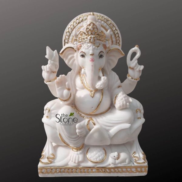 magnificent, highly polished white marble statue of Lord Ganesha, depicted seated in a relaxed yet regal posture, against a solid dark gray background. The pristine white marble gleams softly, beautifully accented by intricate gold detailing that emphasizes the sculpture's fine craftsmanship and traditional Indian artistry. Ganesha is rendered with his characteristic elephant head, large, fan-like ears, and a single tusk, symbolizing his wisdom and ability to overcome duality. His trunk curves gently to his left, and a small red tilak mark adorns his forehead above his serene, dark eyes. He wears an elaborate, multi-tiered gilded crown with delicate carvings and is backed by a similarly ornate, golden aureole or prabhamandala, which frames his head. His four arms each hold symbolic items crucial to his iconography: the upper right hand grasps a paasha (noose), representing control over desires and attachments, while the upper left holds an ankusha (goad), symbolizing guidance and spiritual awakening. His lower right hand is raised in the Abhaya Mudra, a gesture of fearlessness, blessings, and protection, with the palm facing forward. His lower left hand gently cradles a small modak (sweet dumpling), Ganesha's favorite treat, symbolizing the rewards of spiritual pursuit and the sweetness of liberation. Gold bangles adorn his wrists, and a richly gilded necklace drapes around his plump belly, a symbol of prosperity and abundance. The gold trim also highlights the flowing drapery of his dhoti, adding to the idol's luxurious feel. He is seated comfortably on a multi-tiered, intricately carved base, also in white marble with complementary gold accents. At the lower left of the base, his loyal vahan (vehicle), a small mouse named Mushika, is sculpted, appearing to gaze up at his Lord. The subtle branding logo, "the Stone Studio," is visible on the lower left side of Ganesha's body, indicating the origin of this finely crafted religious sculpture. This exquisite Ganesha murti, with its luxurious gold leaf embellishments and smooth, detailed finish, embodies traditional Hindu devotional art, perfect for spiritual practice, home altars, or as a decorative piece symbolizing good fortune, wisdom, and the removal of obstacles in a vibrant cultural setting.