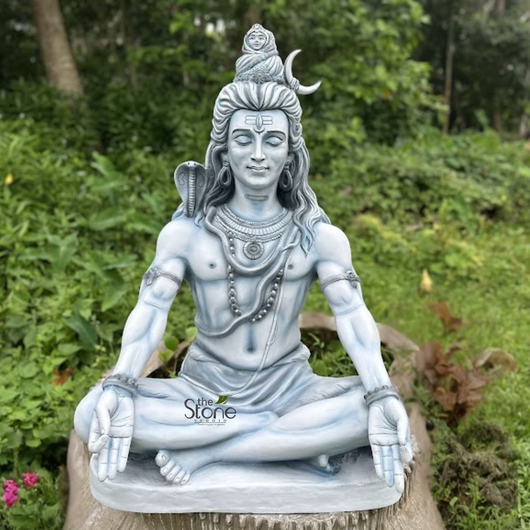 Shiv ji Murti 3ft 2 majestic and serene statue of Lord Shiva, the prominent Hindu deity, is captured in this image, seated in a meditative lotus position (Padmasana). His hands are gently positioned in his lap, palms open and stacked in a Dhyana Mudra, symbolizing profound contemplation and inner peace. The statue is crafted from a light grey, almost off-white material with subtle blueish undertones, suggesting it is made of polished stone, resin, or concrete, giving it an ethereal and tranquil appearance. Shiva's face bears a peaceful, contemplative expression, with his eyes serenely closed. His forehead is marked with the traditional Tripundra (three horizontal lines), symbolizing his divine attributes and third eye. His long, flowing Jatamukuta (matted hair) is intricately styled and adorned with a delicate crescent moon (Chandra) on the right side and a small, detailed figure of the goddess Ganga gracefully emerging from his locks at the crown. A stylized serpent (Naga) is coiled and draped gracefully over his left shoulder, while he wears multiple strands of Rudraksha beads as necklaces and armlets, along with a simple dhoti (loincloth). The statue depicts a lean yet powerful physique, emphasizing asceticism and strength. The sculpture is positioned outdoors on a rustic, natural-looking base, resembling a tree stump or a roughly carved stone block. It is surrounded by lush, vibrant green foliage and distant trees, creating a tranquil and spiritual garden or forest setting. A few small pink flowers are visible near the base on the left, adding a touch of color to the verdant background. The soft, natural lighting further enhances the statue's calm and spiritual aura. This detailed Shiva murti (idol) effectively conveys the deity's iconic attributes and his state of deep meditation, making it a powerful visual for articles discussing Hindu spirituality, meditative practices, garden art, or stone craftsmanship related to religious sculptures. A watermark for 'the Stone Studio' is subtly placed in the lower central area of the statue.
