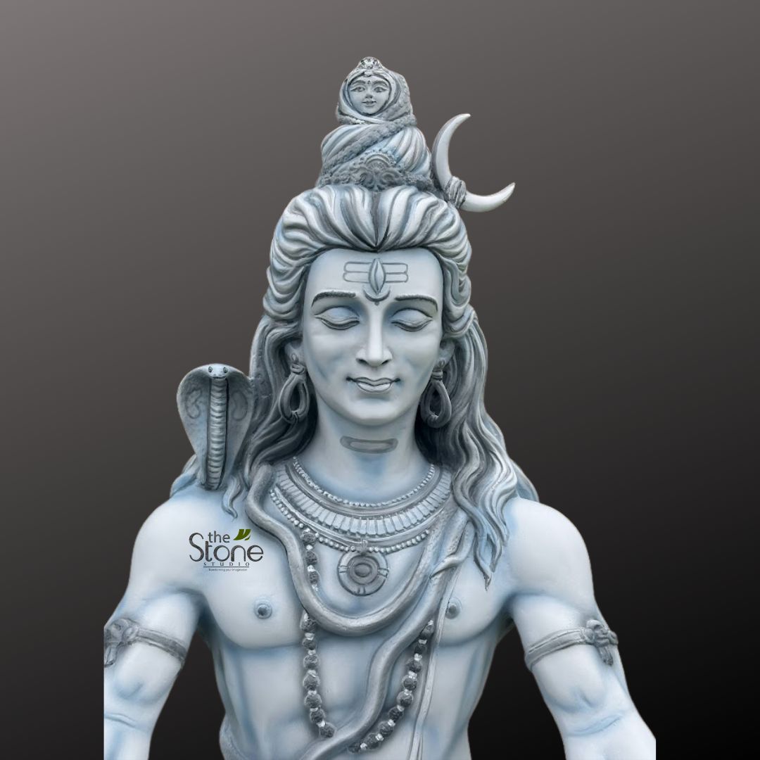 Shiv ji Murti 3ft 5 highly detailed and serene statue of Lord Shiva is depicted, rendered in a striking monochromatic palette of light grey and muted blue tones, suggesting carved stone, marble, or a similar durable material. The sculpture captures Shiva from the chest upwards, presented against a smooth, gradient dark grey background that accentuates its intricate details and spiritual presence. Shiva's face, the central focus of the image, exudes profound peace and tranquility, with his eyes gently closed in deep meditation. A distinct white 'Tripundra' or 'Tilak' mark, symbolizing his third eye and ascetic nature, is prominently visible on his forehead. His features are finely sculpted, with a soft smile gracing his lips and elongated earlobes adorned with simple, circular earrings. His long, matted hair (Jata) is intricately sculpted, flowing down his shoulders and expertly piled high into a majestic topknot (Jata-mukuta). Within the topknot, a small, graceful figure of Goddess Ganga emerges, symbolizing the sacred river's descent through Shiva's hair to the earth. Beside her, a prominent crescent moon (Chandra) adds to his divine iconography, signifying his control over time. Draped around his neck and resting over his right shoulder is a sculpted cobra (Naga, often Vasuki), its hood subtly raised near his ear, representing his mastery over fear, primal forces, and attachment. He wears multiple strands of bead necklaces, including a prominent choker-style piece with a central medallion and a longer mala of beads cascading across his bare, muscular chest. Simple, stylized armlets adorn his upper arms, completing his powerful yet ascetic appearance. The overall texture of the statue appears smooth and polished, highlighting the craftsmanship involved in its creation. A subtle white, stylized watermark "the Stone Studio" is visible on the lower left side, overlapping Shiva's shoulder, indicating the creator or vendor of this exquisite spiritual art piece. This sculpture would be highly relevant for articles discussing Hindu mythology, spiritual practices, the symbolism of Lord Shiva, religious art, or artisan craftsmanship in creating devotional sculptures for home or temple adornment.