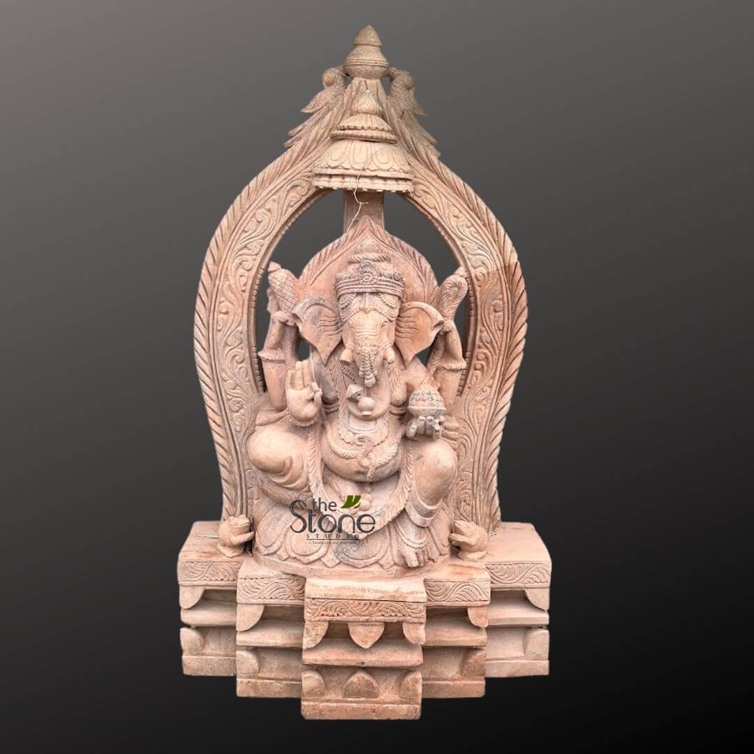 3.5 feet Ganesha Statue 1 majestic, intricately carved stone sculpture of Lord Ganesha, the revered Hindu deity, rendered in a light reddish-brown or sandy-hued stone, likely sandstone. The sculpture depicts Ganesha seated comfortably in a traditional pose, adorned with elaborate ornamentation. He has four arms: the upper right hand holds an axe (parashu), the upper left a noose (pasha), the lower right is raised in the Abhaya Mudra, offering blessings and dispelling fear, and the lower left hand holds a small bowl of modaks (sweet dumplings), symbolizing prosperity and abundance. Ganesha's large elephant head features prominent, fanned ears, a gracefully curved trunk, and a detailed crown. His portly belly is visible, draped with a sacred thread (yajnopavita), and he wears multiple necklaces, armlets, and anklets, all meticulously carved. The deity is framed by an elaborate, tall, arched Prabhavali (aura or archway) that rises behind him. This archway is richly carved with traditional Indian motifs, including stylized foliage, floral patterns, and possibly mythical creatures or celestial figures along its intricate borders. The apex of the arch features an ornate finial, resembling an umbrella or miniature temple tower, flanked by decorative bird-like elements, adding to the grandeur and divine aura of the piece. The entire sculpture rests on a substantial, multi-tiered rectangular pedestal, also intricately carved with geometric patterns and layered steps, providing a stable and regal foundation. The craftsmanship is highly detailed and precise, showcasing the skill of the artisan in traditional stone carving techniques. The sculpture is presented against a neutral, dark gray background, which highlights the delicate textures, three-dimensional depth, and the natural color of the stone. A discreet watermark "The Stone Studio" is visible on the lower front of the sculpture, suggesting its origin or commercial context, connecting this beautiful Ganesha idol to themes of artisanal stone art, spiritual decor, and custom stone carvings for religious or home ornamentation.
