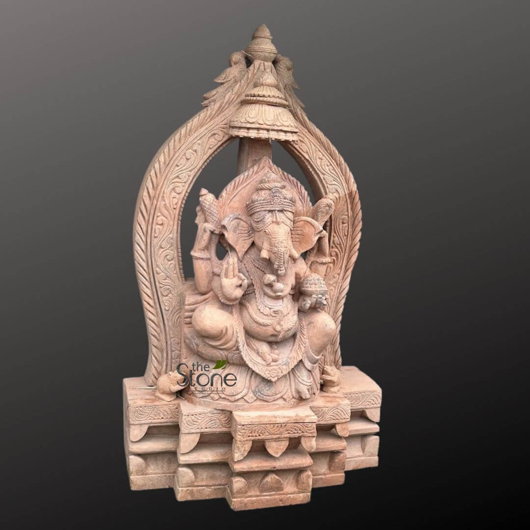 3.5 feet Ganesha Statue 6 majestic, intricately carved sandstone sculpture of Lord Ganesha is depicted against a plain, dark gray background, emphasizing its detail and texture. The Hindu deity Ganesha is seated in a regal and serene posture, likely a variation of Padmasana (lotus position), with his pot-bellied torso adorned with finely carved necklaces, armlets, and a sacred thread (yagnopavita). His iconic elephant head features large, prominent ears, gentle eyes, and a trunk gracefully curled towards his left, reaching down towards a small bowl of modaks (sweet dumplings) held in his lower right hand. One of his tusks is visibly shorter, a common iconographic representation. Ganesha has four arms, each holding significant attributes: his upper right hand clasps an ankusha (goad), while his upper left holds a pasha (noose). His lower left hand is raised in abhaya mudra, a gesture of fearlessness and blessing, with the palm facing outwards. Behind Ganesha, an elaborate arched prabhamandala (aureole or throne back) frames his figure, featuring intricate scrollwork, swirling floral patterns, and stylized mythical creatures or foliage that culminate in a decorative finial at the very top. The entire structure rests on a multi-tiered, stepped pedestal, also adorned with carved geometric patterns and traditional Hindu designs along its edges. To the left of the base, Ganesha's vahana (mount), a tiny mouse named Mushika, is sculpted, looking upwards at the deity. The sculpture's warm, reddish-brown hue and textured finish suggest it is crafted from high-quality natural stone, likely sandstone or a similar earthy material, highlighting the exceptional artistry and craftsmanship involved. A watermark "the Stone Studio" is visible on the lower left, indicating the source of this exquisite piece. This image would be relevant for articles discussing traditional Indian stone carving, Hindu religious art, spiritual home decor, bespoke deity sculptures, or the work of artisan stone studios specializing in cultural and religious iconography.