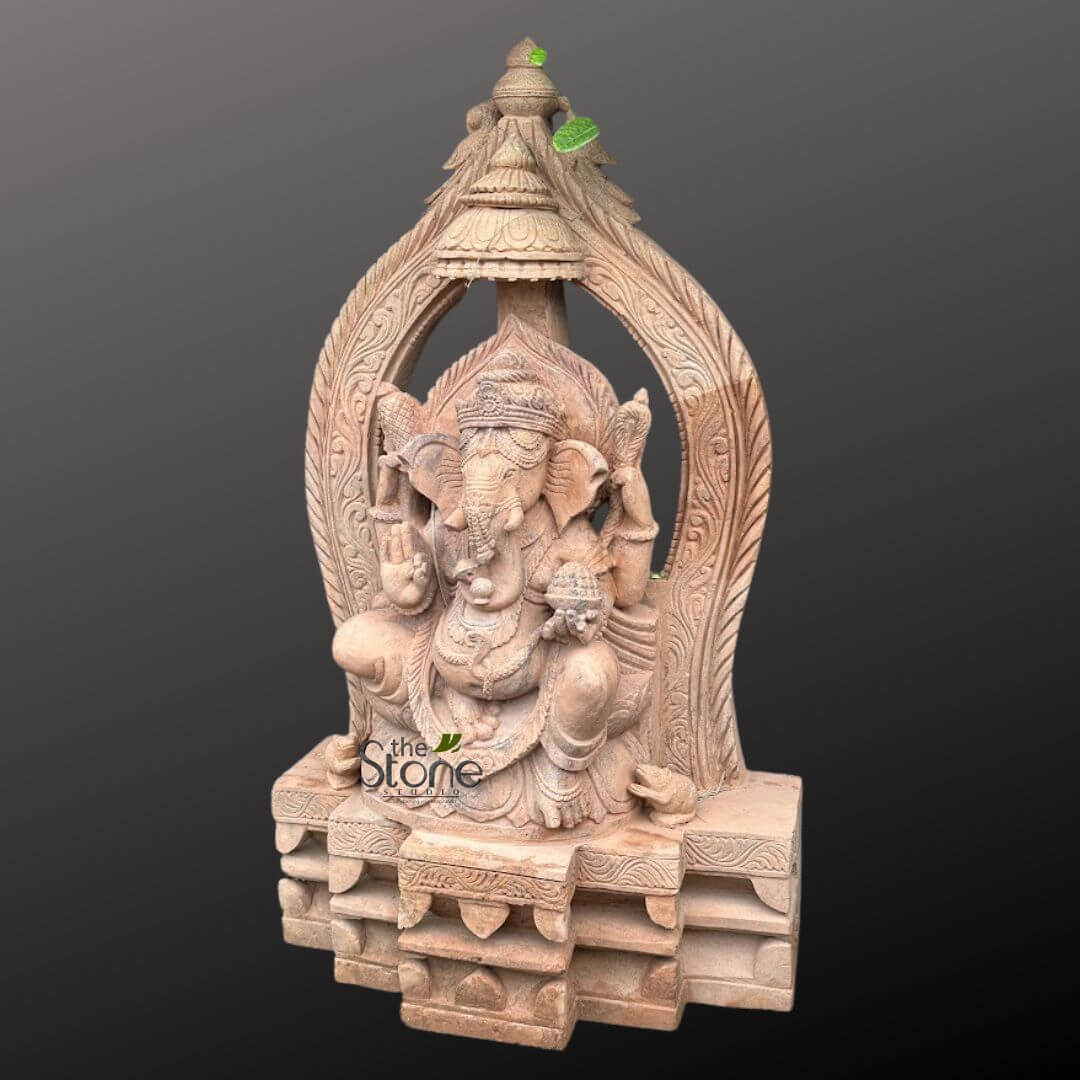 3.5 feet Ganesha Statue 4 magnificent, meticulously hand-carved stone statue of Lord Ganesha, the revered Hindu deity, is presented in a full-view shot against a deep, contrasting grey background that makes the sculpture stand out. This large, traditional Indian Murti is crafted from what appears to be a light brown or sandy-colored stone, possibly sandstone, showcasing its natural texture and the exceptional skill of the artisan. Lord Ganesha is depicted seated gracefully in Lalitasana, a relaxed posture with one leg folded and the other descending, atop an elaborate, multi-tiered pedestal or simhasana. His iconic elephant head features prominent, fanned ears and a trunk elegantly curved towards his left, gently reaching for a modak (sweet dumpling) held in his lower right hand. His four arms are distinctly carved: the upper left hand firmly grasps a parashu (axe), while the upper right holds a pasha (noose). His lower left hand is raised in Abhaya Mudra, a gesture of fearlessness and blessing, inviting prosperity and removing obstacles. The deity is richly adorned with finely detailed traditional jewelry, including an ornate crown, a mala (necklace), armbands, and a sacred thread (yagnopavitam) draped across his chest. His prominent pot belly, a symbol of abundance and prosperity, is also clearly defined. Behind Ganesha, an exquisitely carved prabhavali (arch or aureole) frames the deity, featuring intricate floral and leafy motifs that rise to a decorative finial at the very top, echoing classical Indian temple art and design. At the lower right of the pedestal, Ganesha's faithful vahana, Mushika, the mouse, is subtly depicted near his feet. The entire stone carving exemplifies exceptional craftsmanship, with sharp, precise details evident throughout the idol and its surrounding architectural elements. The visible 'the Stone Studio' watermark on the statue's base suggests its origin from a specialized artisan studio, emphasizing the quality and authenticity of this hand-carved sculpture. This detailed Ganesha statue is a powerful representation of devotion and traditional artistry, making it an ideal piece for spiritual practice, sacred home decor, or as a significant acquisition of traditional Indian sculpture.