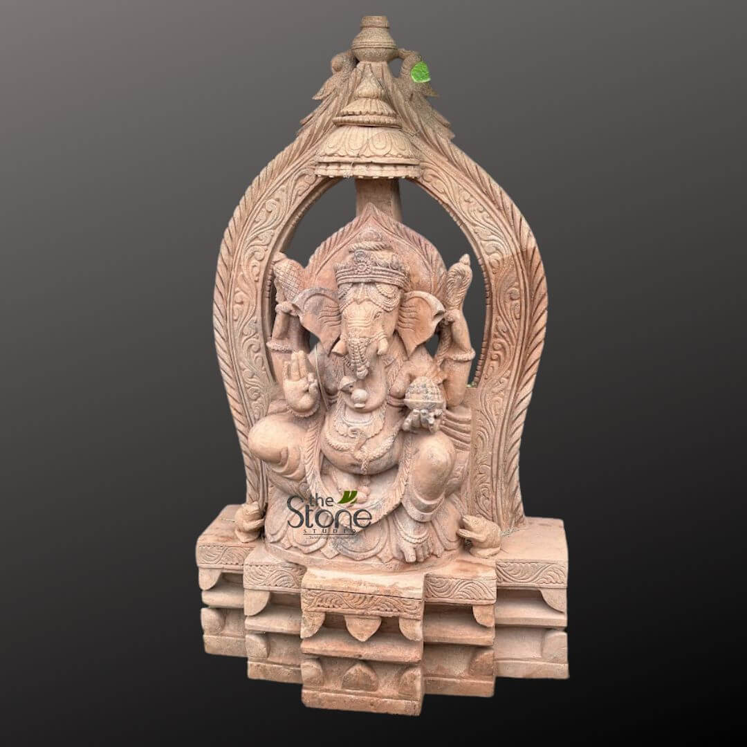 3.5 feet Ganesha Statue 3 magnificent, intricately carved sandstone statue of Lord Ganesha, presented against a plain dark grey background that emphasizes its detailed craftsmanship. The revered Hindu deity is depicted in a serene, seated posture, with his left knee raised and his right leg folded, exuding a sense of calm and wisdom. Ganesha's distinctive elephant head features large, gracefully carved ears, a prominent trunk curled elegantly towards his left, a complete left tusk, and a traditionally broken right tusk. He is adorned with a meticulously sculpted crown (mukuta), ornate necklaces, armlets, and a sacred thread (yajnopavita) draped across his chest. The four-armed figure holds specific attributes: an ankusha (goad) in his upper right hand and a pasha (noose) in his upper left hand. His lower right hand is raised in the 'Abhaya Mudra,' a gesture of fearlessness and blessing, while his lower left hand gently holds a modak (sweet dumpling), towards which his trunk points. Behind Ganesha, an elaborately carved archway, often referred to as a 'prabhavali,' frames his form, showcasing intricate floral, vine, and swirling mythological patterns, culminating in a decorative finial at the very top. The statue rests on a sturdy, multi-tiered, rectangular pedestal, also carved with geometric and architectural details that complement the main figure's design. Made from a beautiful light reddish-brown or buff-colored sandstone, the statue's warm, earthy tones highlight the exquisite craftsmanship and the natural texture of the stone. This exceptional piece of traditional Indian sculpture is rich in cultural and spiritual symbolism, embodying strength, wisdom, and good fortune, making it a significant focal point for devotion, interior decor, or as a timeless representation of spiritual artistry and master craftsmanship in natural stone.