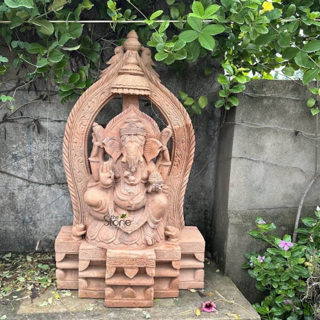 3.5 feet Ganesha Statue 2 magnificent, intricately hand-carved sandstone statue of Lord Ganesha, depicted in a seated posture, dominates the center of this outdoor scene. The statue exhibits a warm reddish-brown, almost terracotta-like hue, characteristic of finely worked stone. Ganesha's elephant head is clearly visible with large, prominent ears, a single tusk (his right), and an elegantly curving trunk. He wears an ornate, multi-tiered crown and is adorned with various necklaces, armlets, and a waistband, all meticulously sculpted. His four arms hold traditional attributes: his upper right hand holds what appears to be an axe or goad (parashu/ankusha), his upper left hand grasps a noose (pasha), while his lower right hand delicately holds a small bowl filled with modaks, his favorite sweet. His lower left hand is raised in the Abhaya Mudra, a gesture symbolizing blessing, fearlessness, and protection. Ganesha is seated upon a lotus-style pedestal, which is itself part of a larger, multi-tiered rectangular base. This base features architectural detailing with recessed panels and subtle decorative motifs. In front of the main base, a smaller, single-tier stone block serves as an offering platform. Behind the deity, an exquisitely carved, arch-shaped prabhavali (aureole or frame) frames Ganesha, showcasing delicate scrollwork, floral patterns, and is topped with a decorative finial. The entire stone carving is set outdoors against a textured, light grey concrete wall, which appears slightly weathered. Lush, vibrant green foliage and climbing vines gracefully drape over the top of the wall and down its sides, forming a natural canopy and backdrop for the statue. To the right, a small plant with delicate purple and pink flowers (resembling periwinkles or vinca) adds a touch of natural color. The ground beneath the statue is a flat concrete surface with a scattering of dry leaves and a single fallen purple blossom, completing the serene, spiritual garden or courtyard aesthetic. The watermark "the Stone" is subtly visible on Ganesha's belly. This detailed piece exemplifies traditional Indian religious art and artisanal stone carving, connecting to themes of spirituality, Hindu deity worship, and bespoke decor for homes or temples.