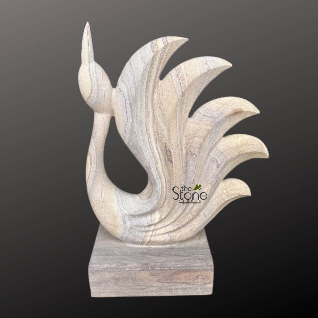 Abstract Peacock Statue 2ft 6 striking and elegantly carved stone sculpture, likely made of sandstone or a similar variegated natural stone, stands prominently against a dark charcoal grey background. The artwork depicts a highly stylized, abstract rendition of a bird, possibly a peacock or a graceful swan, in a dynamic and upward-reaching pose. The sculpture's form is fluid and organic, starting with a slender, pointed head and beak that extends upwards from a gracefully curving neck. The body flows seamlessly into a magnificent, fanned tail composed of five distinct, undulating plume-like segments. These segments are layered and decrease in size from the top, creating a sense of movement and lightness despite the solid material. Each part of the sculpture, from the elegant curve of the neck to the intricate layering of the tail feathers, showcases smooth, polished surfaces that catch the light gently. The natural stone itself is a key visual element, featuring beautiful, organic striations and veins in a palette of soft, earthy tones. Predominantly, there are creamy off-whites, warm beiges, and various shades of soft grey, interspersed with delicate hints of light brown or even subtle purplish-pink hues that swirl and flow across the surface of the sculpture. These natural patterns highlight the geological beauty of the material and give the piece a unique character. The entire artwork rests on a sturdy, rectangular block base, also crafted from the same variegated stone, providing a solid foundation for the ascending form. The dark, uncluttered background ensures the light-toned sculpture stands out sharply, allowing its intricate details, smooth curves, and the natural beauty of the stone's patterns to be fully appreciated. A subtle "the Stone Studio" logo is visible in the bottom right corner, identifying the artisan or workshop behind this exquisite handcrafted piece. This sculpture is a superb example of contemporary stone artistry, ideal for illustrating articles on modern minimalist design, luxury home decor, custom stone carvings, or the integration of natural materials into high-end artistic expressions.