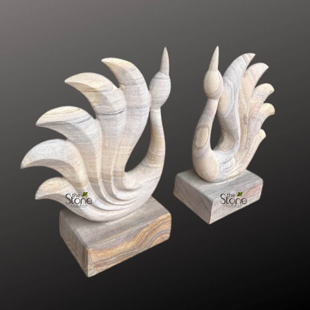 Abstract Peacock Statue 2ft 5 striking image features two meticulously hand-carved natural stone sculptures, crafted from layered sandstone, showcasing a beautiful interplay of natural colors and elegant forms. Each piece depicts a highly stylized, elegant bird, reminiscent of a peacock or swan, with a gracefully arching neck culminating in a pointed head, and a distinct, fanned-out tail. The larger sculpture, positioned prominently on the left foreground, displays five sweeping, distinct feather-like elements forming its tail, which emanate from a smooth, curving body. Its surface reveals intricate horizontal and wavy striations in soft beige, creamy white, pale grey, and subtle hints of warm ochre or light brown, characteristic of sedimentary rock formations. This natural patterning adds depth and organic texture to the smooth, polished curves of the stone artwork. It rests securely on a rectangular plinth of matching striped sandstone, providing a stable foundation and completing the cohesive aesthetic. Slightly behind and to the right, a smaller, equally elegant sculpture mirrors the design, featuring a similar slender profile and four prominent fanned elements that suggest a bird in display. Its stone also exhibits the same captivating natural veins and earthy tones, though its patterns might differ slightly given the unique nature of each natural stone carving. Both sculptures embody fine artisan craftsmanship, transforming raw material into graceful, modern art pieces. These decorative stone carvings would serve as sophisticated accent pieces for interior design, suitable for living rooms, studies, or office spaces, reflecting a blend of natural beauty and contemporary artistic expression. The minimalist dark grey background allows the natural hues and forms of the sandstone sculptures to stand out, while the "The Stone Studio" logo is subtly visible on the bases of both objects, identifying the maker of these unique stone artifacts.