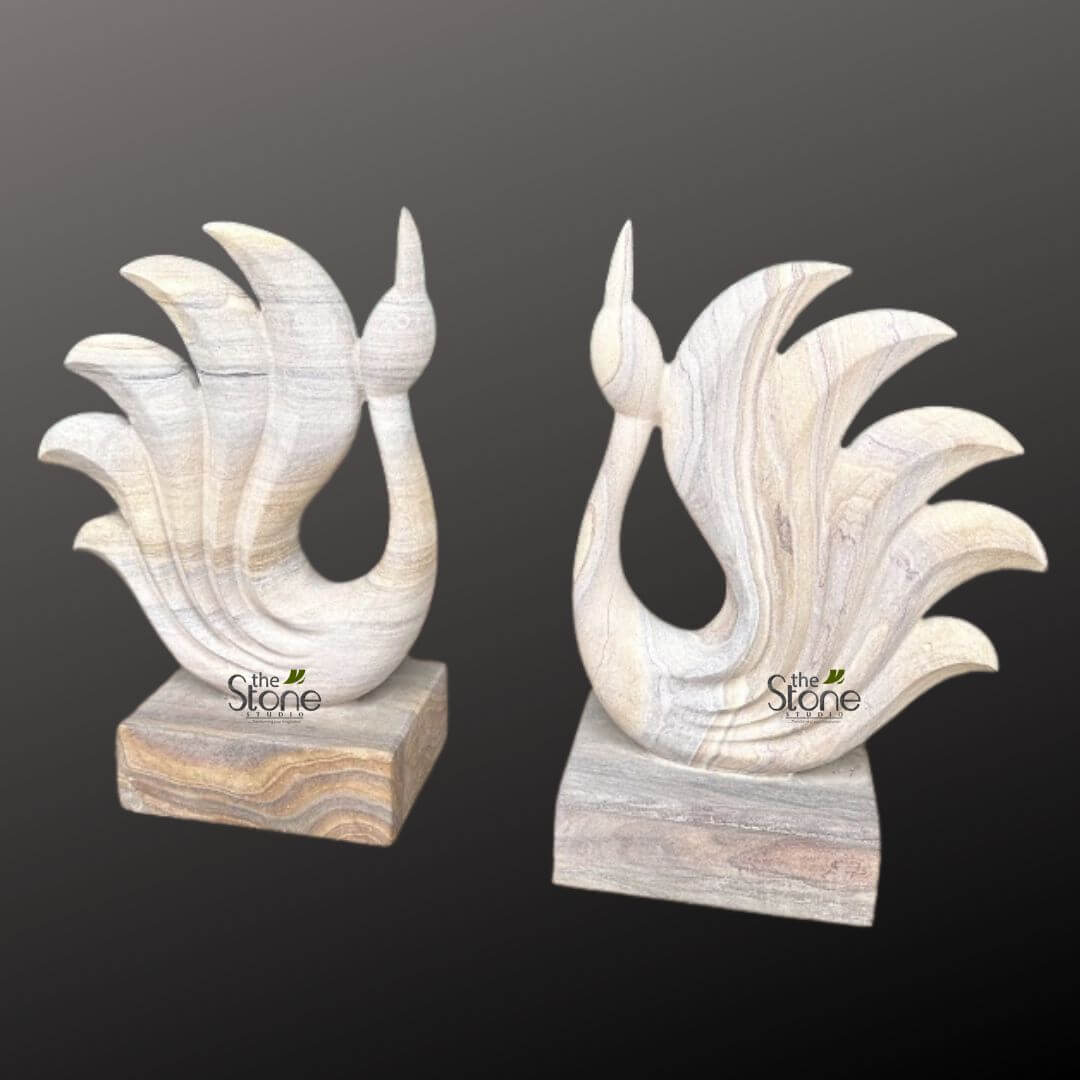 Abstract Peacock Statue 2ft 4 striking pair of hand-carved natural stone sculptures, presented against a minimalist dark grey to black background, showcasing intricate craftsmanship and the inherent beauty of the material. Each sculpture is crafted from a light-colored, variegated stone, likely sandstone or marble, exhibiting beautiful natural veining in shades of cream, beige, soft grey, and subtle browns that run vertically through the bird's form and horizontally across its base. Both pieces depict highly stylized, graceful birds, reminiscent of elegant swans or peacocks. The birds feature an elongated, slender neck curving upwards to a delicate, pointed head or beak. The body of each bird gracefully flows into a magnificent, fanned tail composed of five distinct, fluidly carved elements that fan outwards, suggesting feathers or plumage. The sculpture on the left appears slightly smaller and has warmer, more beige and brown tones in its base, while the sculpture on the right is marginally taller and features a base with cooler grey and white horizontal banding. Each sculpture rests securely on a rectangular, solid block base made from the same natural stone, providing stability and complementing the artistic forms above. A discreet "the Stone STUDIO" logo is visible in white and green on the front surface of the base for both sculptures. These exquisite stone art pieces highlight masterful stone carving techniques and the unique patterns found in natural materials, making them ideal decorative accents or luxury home accessories, perfect for modern or contemporary interior design settings.