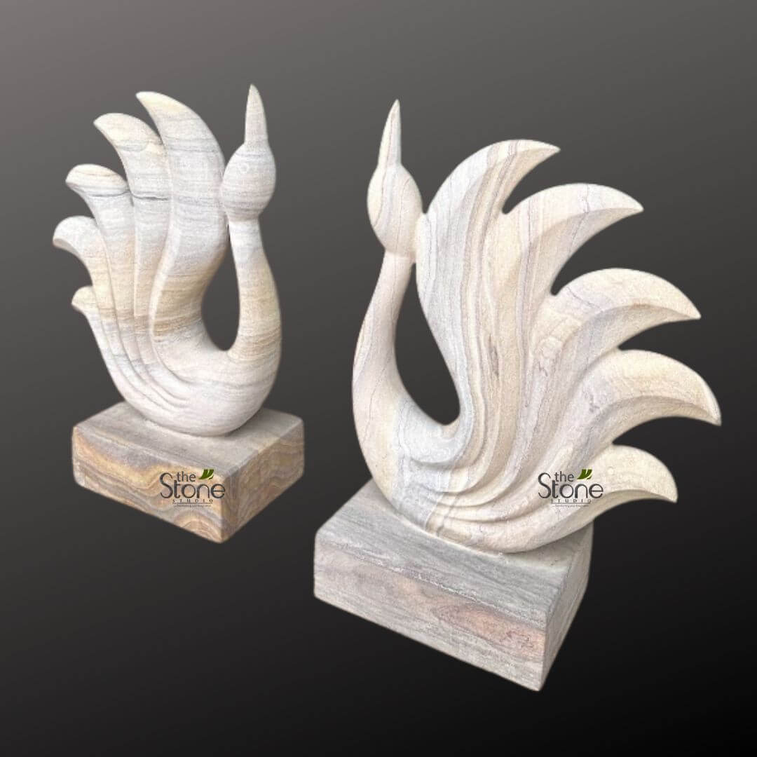 Abstract Peacock Statue 2ft 3 This image features two elegant, handcrafted stone sculptures, presented as a harmonious pair, against a plain, dark grey background that accentuates their light coloration and intricate forms. Each sculpture depicts a stylized bird, possibly a majestic peacock or a graceful swan, rendered in a minimalist yet artistic fashion, with gracefully curved lines and smooth contours. The birds are carved from a beautiful, light-colored natural stone, likely a type of sandstone or marble, characterized by prominent horizontal and diagonal striations in shades of cream, beige, light grey, and subtle hints of warm brown. This natural veining provides a unique, organic texture and visual depth to the polished surface of each piece, highlighting the raw beauty of the stone material. Each bird features a slender, upward-curving neck culminating in a refined, pointed head. The body flows seamlessly into a magnificent, fanned tail composed of five distinct, rounded, feather-like segments that spread upwards and outwards, creating a dynamic sense of movement and artistic flourish. The edges of these "feathers" are smoothly sculpted, contributing to the overall fluid and sophisticated aesthetic. Both sculptures stand firmly on a solid, rectangular base made from the same naturally veined stone, which adds to their stability and perceived value as premium decor items. The two sculptures are arranged side-by-side, mirroring each other: the left sculpture's bird faces right, and the right sculpture's bird faces left, creating a balanced and symmetrical display. The "The Stone Studio" logo, featuring a stylized leaf graphic, is subtly visible on the front face of each stone base. This detailed image effectively showcases the exquisite craftsmanship and timeless beauty of natural stone decor, making it ideal for articles focusing on luxury home accessories, unique handcrafted gifts, interior design trends, artisan stone carving, or the aesthetic appeal of contemporary abstract animal sculptures that add a touch of nature and sophistication to any modern or traditional living space. The interplay of light and shadow on the sculpted surfaces emphasizes the three-dimensional form and the natural variations within the stone itself.