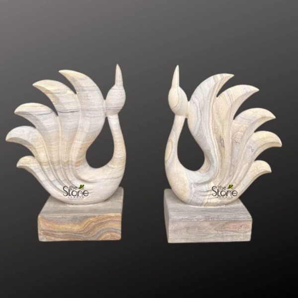 This image displays a magnificent pair of handcrafted stone sculptures, expertly carved to resemble elegant, abstract birds, possibly swans or peacocks, designed to serve as sophisticated decorative accents for any interior space. The two identical yet distinct pieces are presented against a deep, muted charcoal gray background, which allows their natural colors and textures to stand out prominently. Each sculpture features a gracefully curved neck that tapers into a delicate, pointed head or crest, conveying a sense of poise and serenity. The body of the bird flows smoothly into a dramatic, fanned tail composed of five distinct, layered plumes that arc upwards and outwards, giving the impression of majestic wings or a flourishing tail display. The entire form of each bird is sculpted with fluid lines and soft curves. The material used is a stunning natural stone, likely a type of sandstone or layered quartzite, showcasing remarkable horizontal banding or veining. These natural striations are a dominant visual element, appearing in a sophisticated palette of creamy off-white, light beige, and soft gray tones. The veining is skillfully integrated into the carving, following the contours of the bird's body and fanned tail, enhancing the organic feel and highlighting the craftsmanship. The light colors and natural patterns make these pieces versatile for various home decor styles, from rustic to contemporary. Each bird stands securely on a solid, rectangular stone base. The base of the left sculpture features slightly warmer, more pronounced earthy tones with prominent horizontal banding in browns and tans, providing a subtle contrast and grounding the lighter-colored bird. The base on the right sculpture exhibits similar layered stone characteristics, aligning with the overall natural aesthetic. A subtle watermark, "the Stone STUDIO," is visible towards the bottom of each sculpture, identifying the artisan or brand. These exquisite stone bird sculptures are perfect examples of artisanal stone carving, ideal for a mantelpiece, console table, or office desk, reflecting the beauty of natural materials and skilled craftsmanship in decorative art.