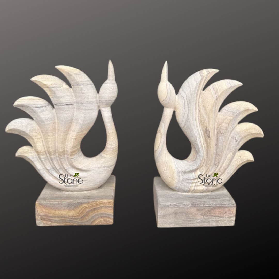 Abstract Peacock Statue 2ft 1 This image displays a magnificent pair of handcrafted stone sculptures, expertly carved to resemble elegant, abstract birds, possibly swans or peacocks, designed to serve as sophisticated decorative accents for any interior space. The two identical yet distinct pieces are presented against a deep, muted charcoal gray background, which allows their natural colors and textures to stand out prominently. Each sculpture features a gracefully curved neck that tapers into a delicate, pointed head or crest, conveying a sense of poise and serenity. The body of the bird flows smoothly into a dramatic, fanned tail composed of five distinct, layered plumes that arc upwards and outwards, giving the impression of majestic wings or a flourishing tail display. The entire form of each bird is sculpted with fluid lines and soft curves. The material used is a stunning natural stone, likely a type of sandstone or layered quartzite, showcasing remarkable horizontal banding or veining. These natural striations are a dominant visual element, appearing in a sophisticated palette of creamy off-white, light beige, and soft gray tones. The veining is skillfully integrated into the carving, following the contours of the bird's body and fanned tail, enhancing the organic feel and highlighting the craftsmanship. The light colors and natural patterns make these pieces versatile for various home decor styles, from rustic to contemporary. Each bird stands securely on a solid, rectangular stone base. The base of the left sculpture features slightly warmer, more pronounced earthy tones with prominent horizontal banding in browns and tans, providing a subtle contrast and grounding the lighter-colored bird. The base on the right sculpture exhibits similar layered stone characteristics, aligning with the overall natural aesthetic. A subtle watermark, "the Stone STUDIO," is visible towards the bottom of each sculpture, identifying the artisan or brand. These exquisite stone bird sculptures are perfect examples of artisanal stone carving, ideal for a mantelpiece, console table, or office desk, reflecting the beauty of natural materials and skilled craftsmanship in decorative art.