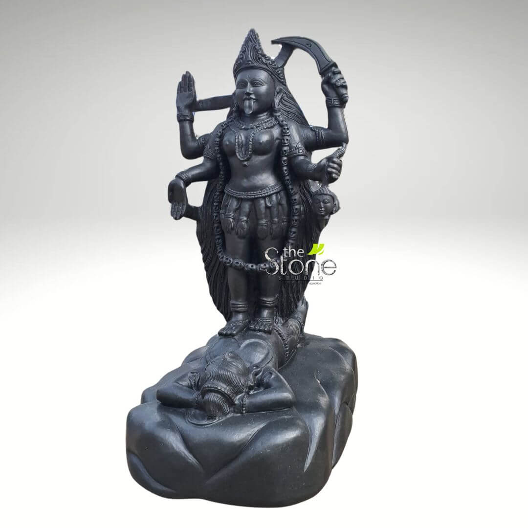 Dakshineswar Maa Kali Statue 27" 6 majestic, dark black stone sculpture of the Hindu goddess Kali stands prominently against a plain, light grey background. The goddess is depicted with multiple arms, at least eight visible, radiating outwards from her torso. She wears an elaborate, tiered crown and has a fierce expression, with wide eyes and her characteristic long tongue protruding from her mouth. Around her neck is a striking garland of severed heads or skulls, and she is adorned with traditional ornaments including necklaces, armlets, and bangles. Her lower garment appears to be a skirt fashioned from severed hands or arms, further emphasizing her formidable nature. In her upper right hand, Kali wields a crescent-shaped scimitar or falchion (known as a khadga), symbolizing the destruction of ignorance and evil. Her upper left hand is raised in the abhaya mudra, a gesture of fearlessness and reassurance to her devotees. Another right hand holds a freshly severed head by the hair, while other hands hold various implements or components of her traditional iconography, possibly a noose (pasha) or a skull cup (kapala). Kali stands triumphantly on the supine body of a male figure, typically identified as Lord Shiva, who lies calmly on his back with his head resting peacefully on his hand. Shiva's serene posture provides a striking contrast to Kali's dynamic and fierce stance. Both figures rest upon a rough-hewn, naturalistic stone base, suggesting a primordial or mountainous landscape. The entire sculpture is rendered in a dark, polished stone, likely granite or basalt, which highlights the intricate carving details and the exceptional craftsmanship. The dark, almost monochrome aesthetic emphasizes the powerful, transformative, and often fearsome aspects of Kali, embodying her role in the destruction of evil and the liberation of souls. This detailed statue serves as a significant representation of Hindu deities, perfect for spiritual devotion, temple adornment, or as a centerpiece in a collection of traditional Indian art. A faint watermark for "the Stone Studio" is visible on the goddess's torso, indicating the origin of this handcrafted stone deity and highlighting the artistry involved in its creation.