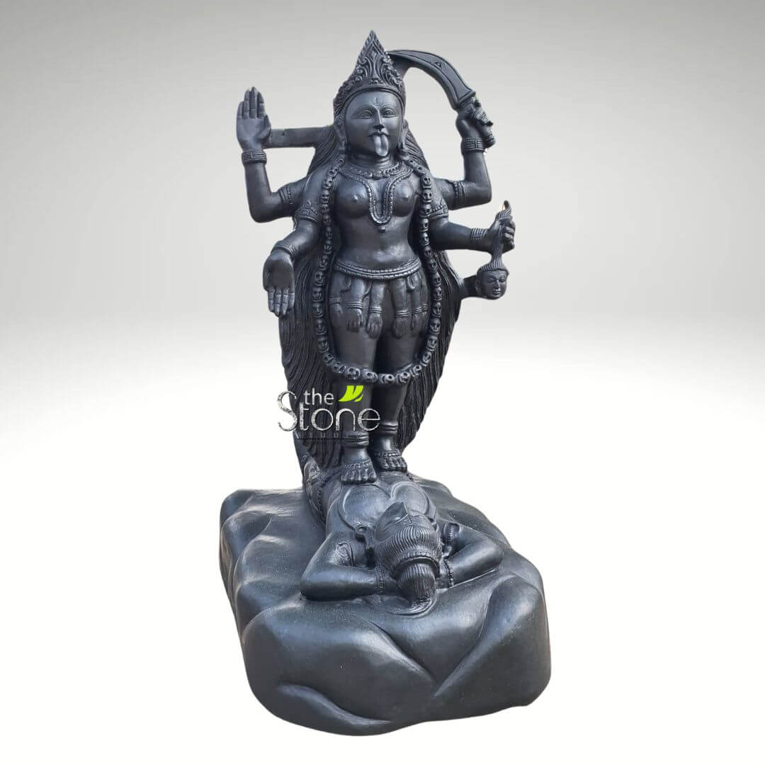 Dakshineswar Maa Kali Statue 27" 5 striking, tall sculpture, meticulously carved from dark, polished black stone, likely granite or marble, depicts the formidable Hindu goddess Kali standing powerfully over the supine figure of Lord Shiva. Kali is portrayed with eight dynamic arms, each expertly sculpted, signifying her immense power. Her fierce visage features a prominent crown, wide, intense eyes, and her iconic long, red tongue protruding from her mouth, symbolizing her insatiable hunger for evil and liberation. Around her neck, she wears a long garland of intricately carved skulls, and her waist is adorned with a skirt-like garment made from severed human arms, emphasizing her destructive and transformative aspects. In her various hands, she wields traditional implements associated with her divine power: a curved scimitar or sword, a trident, a freshly severed head dripping blood, and a bowl (possibly a kapala, for blood or offerings), while other hands display protective and granting mudras (symbolic hand gestures). Her right foot is slightly lifted, while her left foot is firmly planted on the chest of Lord Shiva, who lies serenely on his back on a rugged, rock-like base. Shiva is depicted with a peaceful, calm expression, his eyes closed in deep meditation, hands gently clasped over his stomach, symbolizing his role as the transcendent, unmoving consciousness in the face of Kali's furious cosmic dance. The profound contrast between Kali's dynamic, powerful stance and Shiva's tranquil repose is a central motif in this traditional iconography, representing the interplay of primordial energy (Shakti) and pure consciousness. The intricate details of the deity's jewelry, flowing hair, and the textured base highlight the exceptional craftsmanship of this handcrafted idol, making it a significant piece for spiritual devotion, a temple centerpiece, or an impressive addition to a collection of Indian religious art and sacred sculptures. This detailed depiction effectively conveys the complex symbolism of Kali as both a destructive force against evil and a protective mother leading devotees to liberation.