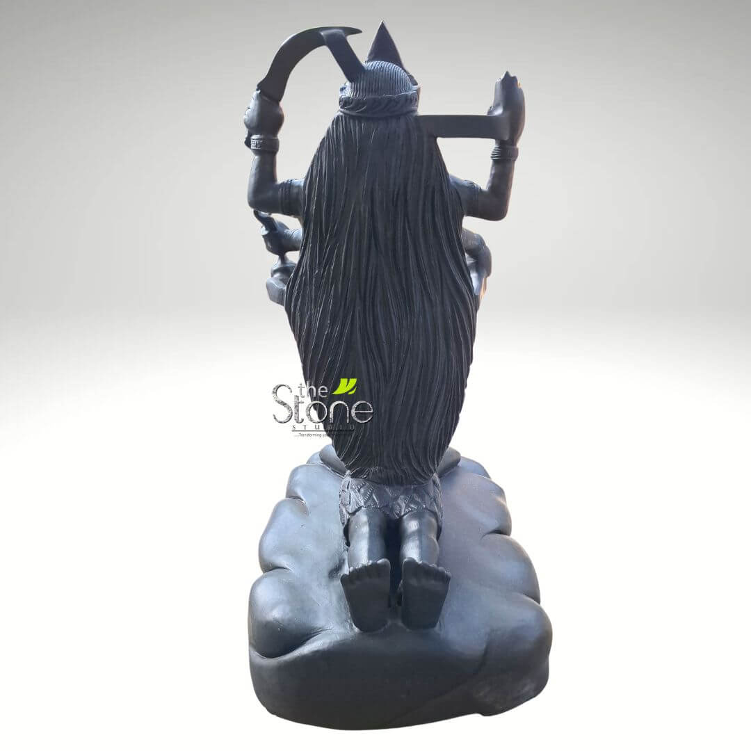 Dakshineswar Maa Kali Statue 27" 4 detailed, large, dark black stone sculpture depicts a powerful, multi-armed Hindu deity, viewed from the back against a plain, light grey gradient background. The craftsmanship suggests it's made from high-quality black granite or a similar dark, durable stone. The deity's most striking feature is her extremely long, flowing black hair, intricately carved to show individual strands, which cascades down her back, covering much of her torso and reaching nearly to her knees. She wears a high, conical crown or headdress, suggesting royal or divine status, possibly adorned with a subtle design or crescent moon at its peak. Four prominent arms are visible, extending outwards from her back. In her upper right hand, she wields a sharp, curved blade, characteristic of a khadga or large sickle, poised as if in motion. Her upper left hand holds a heavy, blunt implement resembling a hammer or an axe head. The lower two arms are bent at the elbows, holding less discernible objects that could be symbolic weapons, ritualistic items, or mudras. The figure's strong, muscular back and shoulders are subtly defined beneath the heavy hair. Around her waist, she wears a textured, short garment or loincloth, possibly depicting animal hide or leaves, adding to her fierce and primal aesthetic. She is depicted kneeling on both knees, with her bare feet visible from behind, heels pointing upwards. The deity rests upon an intricately carved, organic-shaped base that resembles stylized clouds, tumultuous waves, or rocky terrain, enhancing the dynamic and almost ethereal feel of the sculpture. The overall deep, matte black finish of the statue gives it a powerful, ancient, and imposing presence. The watermark "The Stone Studio" is visible on the lower left of the sculpture, indicating the artisan or source. This sculpture strongly evokes imagery of fierce Hindu goddesses like Kali, Chhinnamasta, or a Mahavidya, often associated with cosmic power, time, destruction, and liberation. It serves as a striking example of traditional Indian art and religious iconography, emphasizing the spiritual significance and skilled artistry of stone carving. This detailed depiction would be highly relevant to an article discussing Hindu deities, spiritual art, traditional craftsmanship, or the symbolism of fierce goddesses in mythology.