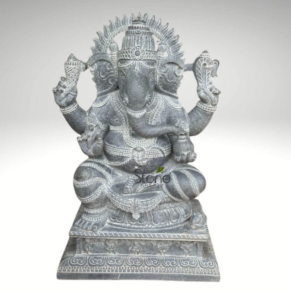majestic, intricately hand-carved stone sculpture of Lord Ganesha is presented centrally against a plain, soft grey studio background. This unique depiction features a multi-headed Ganesha, with a prominent elephant head at the center, flanked by two smaller, human-like heads subtly emerging from either side, looking outwards. The deity is sculpted from dark grey natural stone, possibly granite or a similar durable material, exhibiting a rich, slightly textured surface that highlights its hand-carved nature. Lighter grey or white lines are meticulously etched across the statue, accentuating the elaborate details of his form and attire. Ganesha is seated in a serene, meditative posture, likely Padmasana (lotus pose), with his robust legs crossed. His central elephant head features large, fan-like ears and a benevolent expression. His trunk elegantly curls to his left, appearing to reach towards an offering or sweet (possibly a modak) held in one of his lower hands. He is adorned with a grand, multi-tiered crown (mukuta) on his central head. Behind Ganesha's heads, a magnificent, flame-like or sun-ray halo (prabhamandala) is intricately carved, symbolizing his divine radiance and power. He is depicted with multiple arms, each holding symbolic attributes. In his upper left hand, he cradles a conch shell (shankha), while his upper right hand holds an axe or goad (ankusha). In one of his lower right hands, he prominently displays his broken tusk (Eka Danta), a powerful symbol of sacrifice and wisdom. His corpulent belly is adorned with detailed line carvings representing necklaces, armbands, anklets, a sacred thread (yagnopavitam), and flowing drapery or a dhoti. The statue rests upon a substantial, multi-tiered rectangular pedestal, which is also elaborately carved with continuous patterns of traditional Indian scrollwork, floral motifs, and geometric designs. The meticulous craftsmanship is evident in every detail, from the expressive features to the intricate ornamentation on the base. This exquisite Ganesha statue, a testament to traditional Indian stone sculpting, would serve as an impressive piece of spiritual decor, a focal point for a home altar, a robust garden statue, or a significant addition to any collection of Hindu deity sculptures, naturally blending traditional artistry with devotional symbolism.