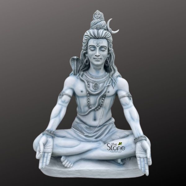 striking, monochromatic statue of Lord Shiva, revered Hindu deity, is depicted in a deeply meditative and serene posture, carved from what appears to be white or light grey stone, marble, or a similar composite material. The detailed sculpture is set against a smooth, dark grey background, making it stand out prominently. Lord Shiva is seated cross-legged (likely Padmasana, the lotus position), with his hands resting open on his knees, palms gently turned upwards in a gesture of contemplation or meditation (Dhyana Mudra). His eyes are softly closed, conveying profound serenity, inner peace, and a sense of deep spiritual absorption, with a subtle, blissful smile gracing his lips. His long, matted hair (Jata) is intricately styled and piled high atop his head in a distinctive topknot, adorned with several significant iconographic elements: a crescent moon (Chandra) is visible on the right side, and a small, distinct figure representing the goddess Ganga is shown flowing gracefully from his topknot, symbolizing the divine river's descent. A prominent third eye (Tilak or Tripundra symbol) is clearly marked on his forehead, signifying wisdom, divine perception, and destruction of evil. A powerful cobra (Naga) is intricately carved, coiled around his left shoulder, with its hood gracefully raised and spread, symbolizing control over ego, danger, and the power of creation and destruction. He wears multiple layered necklaces, including what appear to be Rudraksha beads, and a sacred thread (Yajnopavita) draped across his bare, sculpted torso, which showcases a lean yet muscular physique embodying both asceticism and divine strength. His lower body is modestly draped in a simple, flowing cloth. The craftsmanship is highly detailed, emphasizing the serene expression and the traditional iconography, making this sculpture an embodiment of spiritual strength and tranquility. This depiction of Lord Shiva, often revered as the Adiyogi (first yogi) or Mahadev, would be ideal for spiritual practices, home decor, meditation spaces, or as a focal point for devotion, reflecting themes of enlightenment, self-realization, and the profound journey of yoga.