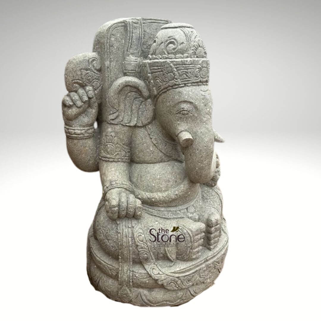 Stone Balinese Ganesh Statue 4ft 6 highly detailed, light gray stone sculpture of Lord Ganesha, the revered Hindu deity with an elephant head, depicted in a dignified and serene seated posture. The statue is meticulously carved from a natural, fine-grained stone, likely sandstone or granite, giving it a nuanced, textured finish that highlights the artisan's craftsmanship. Ganesha's iconic elephant head features a gracefully curved trunk pointing towards the viewer's left. His tranquil eyes are downcast, exuding a sense of peace and wisdom. He wears an elaborate, multi-tiered crown (mukuta) adorned with intricate traditional Indian patterns. Distinctively, the statue portrays Ganesha with a single prominent tusk on his right side (viewer's right), while the left tusk is notably shorter or broken, a classic iconographic detail referencing his role in transcribing the Mahabharata. The robust, pot-bellied form, characteristic of Ganesha, symbolizes prosperity and abundance. He is depicted with four arms: his upper right hand holds a goad (ankusha), while his upper left hand holds a noose (pasha). His lower hands rest gently on his lap; the right hand appears to be holding a sweet delicacy, possibly a modaka, which signifies rewards and spiritual fulfillment. The deity is adorned with finely carved jewelry, including necklaces, armbands (kankanas), and a sacred thread (yajnopavita) draped diagonally across his chest. His draped lower garments are also intricately detailed with traditional motifs and flowing folds. The entire Ganesha figure sits upon an ornate, layered base, which is also richly carved with decorative patterns, suggesting stability and reverence. The sculpture is presented against a plain, neutral light gray background, which effectively isolates the artwork and emphasizes its intricate details and natural stone color. This exquisite Ganesha statue, showcasing traditional Indian artistry and spiritual symbolism, is an ideal piece for spiritual home decor, garden ornamentation, or as a significant cultural art collection item, connecting directly to themes of craftsmanship, Hindu deities, and decorative stone products implied by 'the Stone Studio' watermark.