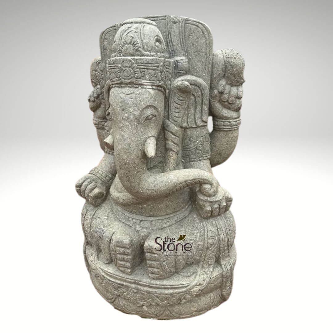 Stone Balinese Ganesh Statue 4ft 5 beautifully detailed and intricately hand-carved natural stone statue of Lord Ganesha, the revered Hindu deity, stands prominently against a plain, soft light gray background, highlighting its texture and form. The statue depicts Ganesha in a seated, meditative pose on an ornate, multi-layered lotus pedestal (Padmasana), symbolizing purity and spiritual awakening. Ganesha, the elephant-headed god, is rendered with distinctive features: his majestic elephant head boasts large, expressive eyes, gracefully prominent ears, and a long, curved trunk that sweeps downwards to the right, appearing to reach for or hold a modak (sweet dumpling), a common offering and symbol of abundance. He wears a meticulously carved crown (mukuta) adorned with traditional patterns, and delicate jewelry including necklaces, armbands, and anklets are etched into his form. The four-armed deity showcases his characteristic implements: the upper two arms are raised, with hands open or possibly holding symbolic items like an ankusha (goad) or pasha (noose), while his lower left hand rests gently on his knee in a gesture of blessing or support. The entire sculpture is crafted from a speckled, light gray natural stone, possibly granite or sandstone, giving it an earthy, rustic yet refined appearance. The rough-hewn texture of the stone adds to its authentic, artisan craftsmanship. Near the base, the subtle logo "the Stone Studio" is visible, indicating its origin as a high-quality product. This elegant stone sculpture serves as an ideal piece for spiritual decor, a home altar, garden embellishment, or as a timeless work of Hindu art, embodying wisdom, prosperity, and the removal of obstacles.