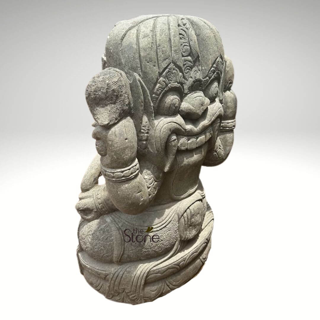 Stone Balinese Ganesh Statue 4ft 4 large, intricately carved grey stone sculpture depicts a formidable, multi-headed mythological figure, possibly a Rakshasa, Yaksha, or a temple guardian from Southeast Asian lore, seated in a meditative or commanding posture. The prominent central head dominates the sculpture, featuring a wide, fierce grin that reveals bared teeth and menacing fangs. Its large, expressive eyes are set beneath a heavily furrowed brow, adorned with ornate, flame-like motifs and a detailed crown-like headdress. Numerous smaller, less distinct faces are subtly incorporated into the sides of the main head and along its crown, creating a powerful, multi-faceted visage that suggests heightened awareness or divine power. The robust figure is depicted with a strong, muscular torso and thick arms adorned with richly detailed armbands or bracelets. Its hands appear to be resting gracefully, possibly in a relaxed mudra or simply placed on its knees, which are part of the figure's integrated base, suggesting a cross-legged seating position. A finely carved sash or garment wraps around its lower body and waist, featuring intricate patterns of swirling foliage and traditional geometric designs, indicative of ancient craftsmanship. Crafted from natural, textured grey stone, the sculpture exhibits a beautifully porous surface that highlights the artisan's skill in traditional stone carving techniques. The varied tones and natural imperfections of the stone add to its authentic, weathered appearance, making it a powerful representation of cultural heritage. This piece embodies the rich artistry found in regions like Thailand, Cambodia, or Bali, making it an ideal visual for articles discussing Asian stone art, mythological figures, ancient spiritual guardians, or cultural home and garden decor. The statue is photographed against a plain, light grey background, allowing its intricate details and imposing form to be the sole focus.