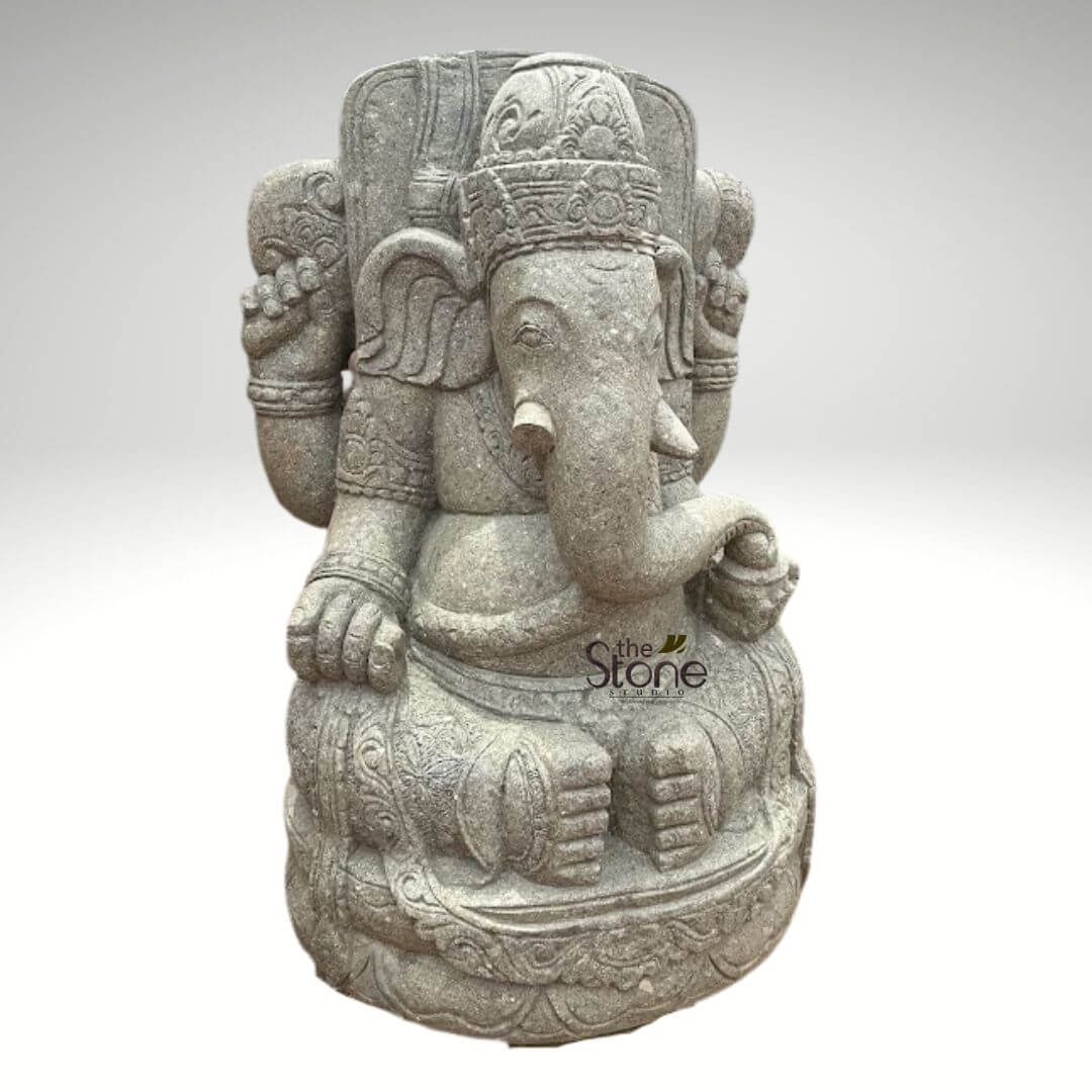Stone Balinese Ganesh Statue 4ft 3 A majestic, intricately carved stone statue of Lord Ganesha, presented front and slightly to the right against a plain, light grey to white background that emphasizes its form and texture. This impressive spiritual art piece is crafted from a speckled, light grey natural stone, likely granite or a similar durable material, giving it a timeless, weathered yet polished aesthetic that highlights its artisanal craftsmanship. Ganesha is depicted in a serene, seated posture, likely Padmasana or a similar meditative cross-legged pose, symbolizing tranquility, wisdom, and steadfastness. He possesses the iconic elephant head with large, gracefully curved ears and a beautifully detailed trunk that curves gently to his left, reaching towards a bowl of sweets in his lower left hand. He has a single intact tusk on his right side, with the left tusk traditionally broken, a symbol of sacrifice and wisdom. Ganesha wears an elaborate, tiered crown (mukuta) with fine, traditional Indian carvings and patterns, and his benevolent face conveys a sense of calm and spiritual presence. The deity is shown with four arms (Chaturbhuja), a common traditional representation emphasizing his divine power and ability to multitask in removing obstacles and bestowing blessings. In his upper right hand, he holds what appears to be an axe or goad (Parashu/Ankusha), symbolizing the cutting of attachments and spiritual guidance. His upper left hand grasps a coiled rope or noose (Pasha), used to capture illusions and worldly desires. His lower right hand gently holds his broken tusk (Danta), which he famously used as a writing instrument for the Mahabharata. And his lower left hand cradles a small, round bowl filled with modak, his favorite sweet, representing spiritual rewards, abundance, and the sweetness of moksha (liberation). Rich, traditional Indian detailing adorns the entire Ganesha sculpture, including intricate patterns on his crown, armbands, necklaces, and the drapes of his clothing. He is seated upon a circular pedestal that features an ornate lotus base or scrollwork, further enhancing its artistic and spiritual appeal. The texture of the stone is visibly granular, showcasing the natural beauty and robust quality of the material. This handcrafted Hindu deity statue, suitable for both indoor and outdoor spiritual decor, a garden statue collection, or as a focal point in a serene space, embodies strength, wisdom, prosperity, and the removal of obstacles. The watermark "the Stone Studio" is subtly placed in the lower right, indicating the source of this exquisite stone carving, suggesting its connection to artisan stone products for home and garden enhancement.