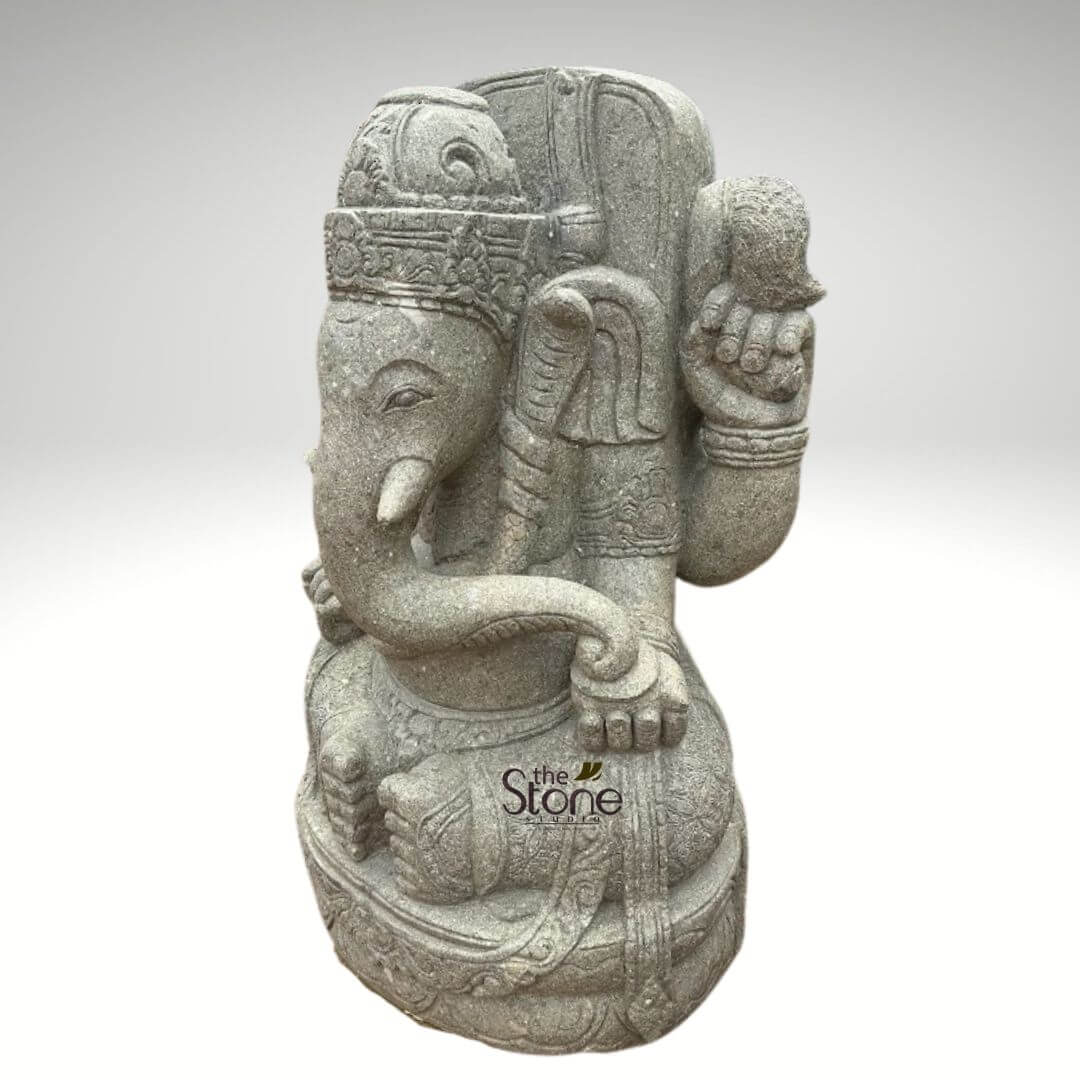 Stone Balinese Ganesh Statue 4ft 2 detailed, majestic grey stone statue of Lord Ganesha, the revered elephant-headed Hindu deity, is shown seated serenely on an intricately carved double-lotus pedestal. The sculpture is crafted from rough-hewn natural grey stone, likely granite or volcanic rock, giving it a rustic yet refined texture with visible grain and subtle variations in tone. Ganesha is depicted with his iconic elephant head, featuring large, rounded ears and a single prominent tusk, while his other tusk is traditionally broken off. His trunk gracefully curves downwards and to the left (from the viewer's perspective). His eyes are carved in a peaceful, half-closed expression, conveying wisdom and calm. He wears an ornate, multi-layered crown adorned with intricate patterns and a subtle flame-like finial at its peak. The deity is shown with four arms, a traditional representation. His front left hand (viewer's right) gently holds what appears to be a small modak (sweet dumpling), a symbol of rewards and blessings. His front right hand (viewer's left) rests on his raised knee, possibly in a gesture of blessing or contemplation. His upper right arm holds an 'ankusha' (goad), and his upper left arm holds a 'pasha' (noose or rope), symbols representing guidance and the control over desires. Elaborate armlets and bangles adorn his wrists and arms, alongside a sacred thread (Yajnopavita) draped across his chest. The double-lotus pedestal he sits upon is a work of art itself, featuring multiple layers of sculpted petals, enhancing the divine and meditative posture. The entire sculpture exhibits robust yet detailed craftsmanship, highlighting the artisanal quality of the hand-carved stone. This spiritual and aesthetic piece is ideal for both outdoor spaces like gardens, patios, or entryways, as well as for indoor spiritual altars or decorative accents. Its durable natural stone construction makes it suitable as a weather-resistant garden ornament or a timeless home decor item. The image is a product photograph, presented against a plain, light grey background, allowing full focus on the intricate details and texture of the statue. A "the Stone Studio" watermark with a leaf motif is subtly visible on the lower right side of the statue, indicating its origin as a handcrafted stone art piece.