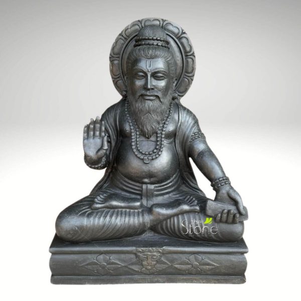 detailed black stone statue of a revered Hindu sage or spiritual master, presented full-front and seated in a meditative cross-legged posture (Padmasana) on an ornate rectangular pedestal. The figure exudes a dignified, serene expression, characterized by a prominent, flowing beard and mustache, and a U-shaped mark (Tilaka or Urdhva Pundra) on the forehead, suggesting devotion. His hair is styled into a neatly coiled topknot, adorned with a decorative headpiece. Behind his head, a large, intricately carved prabhamandala (halo or aura) features delicate lotus petal designs, symbolizing purity and enlightenment. The sage wears simple, flowing robes that drape gracefully over his body, and is adorned with long prayer beads (mala) around his neck, signifying spiritual practice. His right hand is raised in the Abhaya Mudra, a gesture conveying fearlessness, reassurance, and divine protection, while his left hand rests gently on his lap, holding an ancient scripture, book, or palm-leaf manuscript, emphasizing wisdom and knowledge. Carved from dark, polished black granite or basalt, the statue showcases exceptional craftsmanship, with fine details visible in the texture of the beard, the folds of the drapery, and the intricate patterns on the pedestal, which includes floral motifs and a central mythical emblem, possibly a Kirtimukha. This powerful and serene depiction embodies spiritual authority, profound knowledge, and divine grace, making it an ideal representation for articles discussing ancient Indian philosophy, Hindu spiritual traditions, sacred texts, meditation practices, or traditional stone art and sculpture.