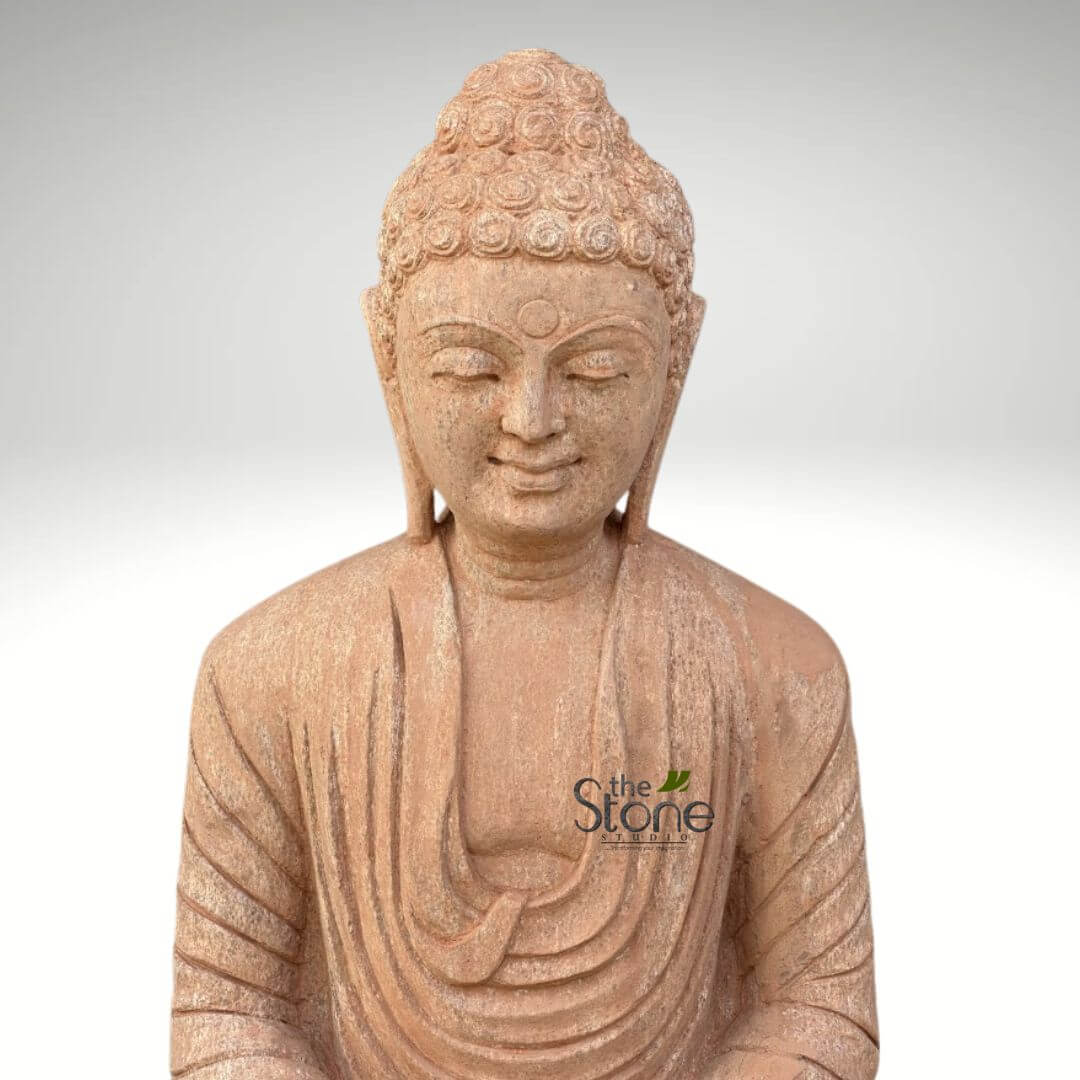 30 inches Garden Buddha Statue 4 meticulously carved stone Buddha statue, presented in a close-up, chest-up view, stands prominently against a clean, light grey gradient background. The statue is crafted from a beautiful reddish-brown sandstone, showcasing a fine, textured finish that highlights the natural beauty of the material. Buddha's face is central, with his eyes gently closed in deep meditation, and a subtle, serene smile gracing his lips, conveying an profound sense of peace, tranquility, and inner harmony. His hair is intricately sculpted into tight, coiled curls that rise to form a distinct ushnisha, the traditional cranial bump symbolizing wisdom. A small, circular urna, a dot symbolizing spiritual insight, is visible between his eyebrows. The drapery of his robes is realistically rendered with flowing, vertical pleats over his left shoulder and chest, adding to the statue's elegant and timeless aesthetic. The overall impression is one of calm reverence, making this handcrafted stone sculpture an ideal piece for spiritual home decor, a peaceful garden setting, or as an artifact representing mindfulness and enlightenment. The logo "the Stone Studio" is subtly visible on the lower left of the statue's chest, indicating its origin as a piece of fine stone carving.