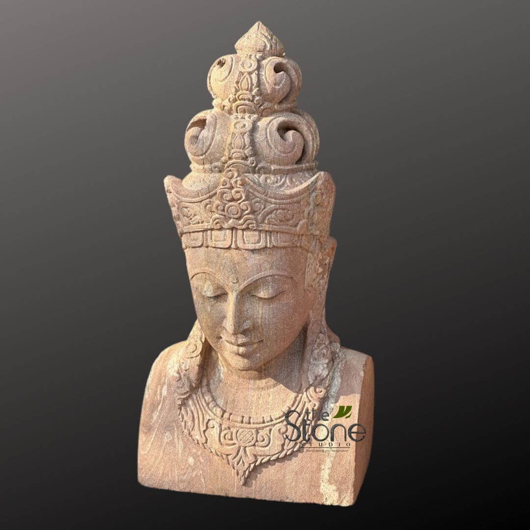 2ft Lord Vishnu Head 6 stunning, finely carved stone bust depicting a spiritual or divine figure, crafted from light reddish-brown or pinkish-hued natural sandstone, showcasing its distinct grain and textured surface. The figure's face exhibits a profound sense of tranquility, with downcast, closed eyes conveying a peaceful, meditative state. Subtle contours define the nose and lips, contributing to the serene expression. Dominating the upper portion is an elaborately tiered and highly ornate crown or ceremonial headdress. It features multiple layers of intricate, swirling patterns, possibly floral motifs, scrollwork, and geometric designs, culminating in a pointed, finial-like apex at the very top. A detailed band with repeating patterns circles the forehead. Below the face, the neck is gracefully carved, adorned with a complex, multi-layered necklace. This sculpted jewelry showcases more delicate patterns, possibly stylized foliage or ancient script-like elements, adding to the figure's regal and revered appearance. The bust extends to include the upper chest and shoulders, with traces of sculpted hair or decorative garlands flowing down the sides, further enhancing its ancient and cultural aesthetic. The craftsmanship highlights traditional carving techniques, making it an exquisite example of spiritual art or ancient sculpture. The earthy, natural tones of the stone contrast subtly with the plain, dark grey background, emphasizing the statue's intricate details and its aura of peaceful contemplation. This piece evokes cultural heritage and spiritual devotion, ideal for discussions on Asian art, stone carving, or meditative practices.