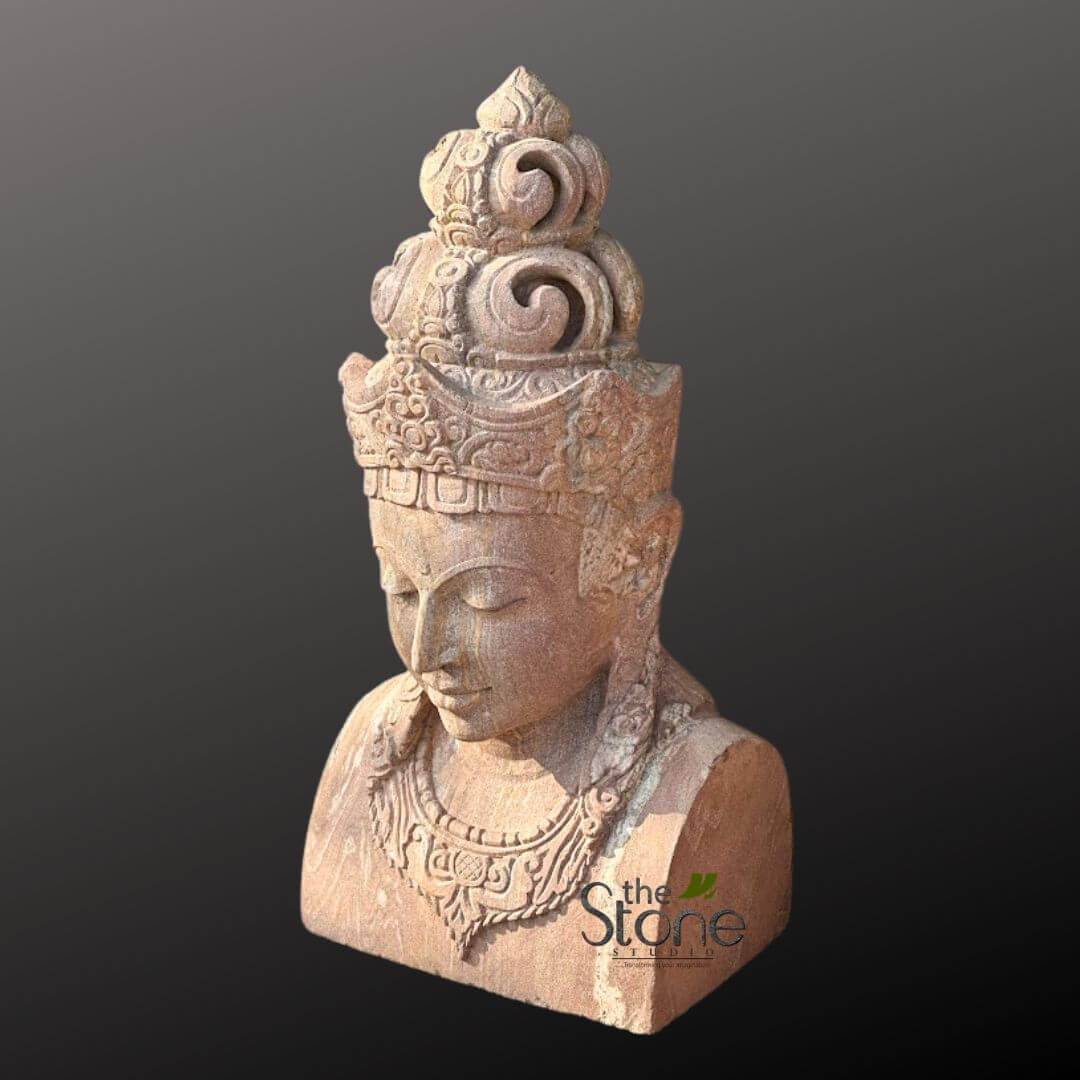 2ft Lord Vishnu Head 5 detailed, eye-level shot of a beautifully hand-carved stone bust, likely made from reddish-brown sandstone, showcasing its natural texture and warm, earthy hues. The sculpture depicts a serene and meditative figure, characteristic of a Bodhisattva or Buddhist deity, with eyes gently closed and gaze directed downwards, conveying a profound sense of inner peace and tranquility. The figure is adorned with an exceptionally intricate and multi-tiered crown. The base of the crown features delicate, finely carved geometric patterns and floral motifs, while the upper tiers are embellished with large, gracefully swirling volutes and decorative scrolls, culminating in a pointed, finial-like apex. Beneath the crown, the figure's face is characterized by a high forehead, a well-defined nose, and soft, closed lips suggesting peaceful contemplation. Elongated earlobes, symbolic of wisdom and spiritual attainment in many Asian traditions, are visible on either side of the head. Around the neck, a magnificent, deeply carved necklace is draped, featuring an elaborate central pendant and symmetrical, flowing patterns that extend over the shoulders, hinting at traditional garments. The shoulders themselves are smoothly sculpted, contributing to the figure's dignified and composed posture. The expert lighting highlights every fine detail and texture of the stone, casting subtle shadows that emphasize the depth of the carving. The bust is presented against a plain, dark grey background, which serves to make the sculpture stand out prominently. A subtle watermark for "The Stone Studio" is visible on the lower right of the bust. This exquisite piece exemplifies traditional stone artistry and cultural heritage, making it an ideal representation for articles on spiritual decor, ancient Asian sculptural techniques, or artisan craftsmanship.