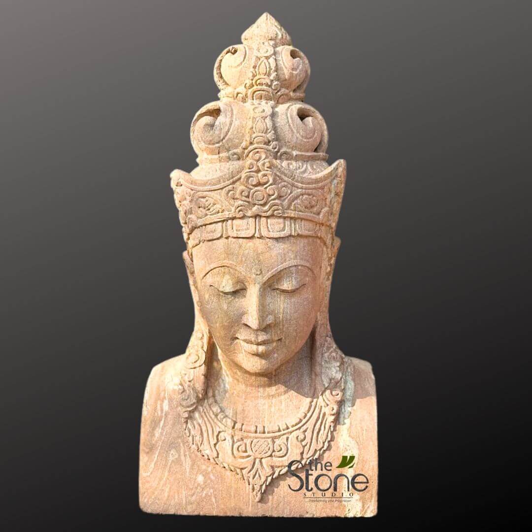2ft Lord Vishnu Head 1 visually striking, full-frontal studio shot of a magnificent hand-carved natural stone bust, evoking a sense of ancient spirituality and artistic craftsmanship. The sculpture, rendered in a warm, light beige to sandy brown natural stone – possibly sandstone, limestone, or volcanic rock – exhibits a finely textured, slightly weathered surface that accentuates its detailed carvings. It represents a serene, idealized spiritual figure, likely a Bodhisattva, Buddha, or a revered deity from Hindu or Buddhist traditions. The figure's face is characterized by an elongated, gentle oval shape, with eyes peacefully closed and downcast in deep meditation, conveying profound tranquility and inner peace. A subtle, ethereal smile graces the lips, and a small urna or 'third eye' dot is visible on the forehead, signifying wisdom and enlightenment. Adorning the head is an exquisitely detailed, multi-tiered crown (mukuta), carved with traditional ornate motifs, including swirling patterns, floral elements, and geometric designs. A majestic spire or finial culminates at the very top, adding to its regal appearance. Stylized hair, resembling elongated strands or dreadlocks, descends gracefully from beneath the crown, resting on the shoulders. The ears are elongated, a characteristic often associated with spiritual figures. A broad, intricately carved necklace or collar encircles the figure's neck and upper chest, mirroring the elaborate patterns of the crown, suggesting a harmonious and complete design. The bust cuts off at the upper chest, showcasing the figure's head, neck, and shoulders. The sculpture is set against a clean, dark grey to black gradient background, which effectively highlights the light-colored stone and allows the intricate details of the craftsmanship to stand out without distraction. A subtle watermark for "the Stone STUDIO" with a leaf motif is visible in the lower right corner, indicating the creator or source of this beautiful, traditional artwork. This piece exemplifies high-quality stone carving and traditional Asian spiritual art, suitable for home decor, garden ornamentation, or as a focal point in a meditation space.