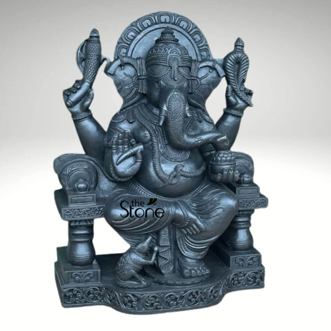 Black Stone Ganesha Sculpture for Garden 3.5ft 6 highly detailed and exquisitely crafted dark grey stone statue of Lord Ganesha, the revered Hindu deity, seated majestically on an ornate throne against a plain light grey background. The sculpture captures Ganesha in a traditional posture, with his iconic elephant head featuring large, fanned ears and a gently curving trunk that reaches towards a small bowl of modaks (sweets) held in his lower left hand. Ganesha is depicted with four arms, symbolizing his divine power. His upper right hand holds an ankusha (goad), while the upper left hand grasps a pasha (noose). His lower right hand is raised in the Abhaya Mudra, a gesture of fearlessness, blessings, and protection, with the palm facing outwards. The deity is richly adorned with an elaborate crown, intricate necklaces, armlets, bracelets, a waist belt, and a prominent sacred thread (yagnopavitra) depicted as a serpent coiled around his large belly. His draped garment (dhoti) shows delicate folds and patterns. At Ganesha's feet, on the richly carved pedestal, sits his loyal vahana (mount), a small, attentive mouse (Mushika), completing the traditional iconography. The throne itself is highly decorative, featuring detailed carvings on its legs and base, and a circular, patterned arch (prabhavali) framing Ganesha's head. The entire piece demonstrates exceptional craftsmanship and attention to detail, characteristic of traditional Indian stone art and sculpture. This magnificent stone idol, likely from "The Stone Studio" as indicated by the watermark, serves as a powerful symbol of wisdom, prosperity, and the remover of obstacles, often used for spiritual devotion, temple adornment, or as an elegant piece of religious decor for homes and gardens.