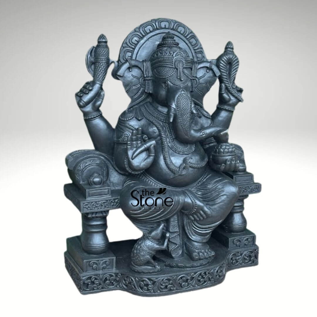 Black Stone Ganesha Sculpture for Garden 3.5ft 5 striking, highly detailed dark charcoal grey Ganesha sculpture, crafted with a rich, textured stone finish, is presented against a plain light grey background. This sacred Hindu deity, revered as the remover of obstacles and god of wisdom, is depicted seated in a regal pose on an elaborately carved, multi-tiered throne. Lord Ganesha's iconic elephant head features large, fanned ears, a gracefully curved trunk that turns to his left, and a single tusk, symbolizing his wisdom and ability to overcome duality. He is adorned with a towering crown, multiple necklaces, armlets, ankle bracelets, and a sacred thread (upavita) draped across his pot-bellied form, all intricately sculpted with fine detail. His four hands each hold symbolic objects: the upper left grasps an ankusha (elephant goad), symbolizing control over negative impulses; the upper right holds a pasha (noose), representing the binding nature of worldly attachments. His lower left hand is raised in the protective Abhaya Mudra, offering blessings and assurance, while his lower right hand holds a bowl brimming with modaks (sweet dumplings), a treat Ganesha is famously fond of. At his left foot, his loyal vahana, Mushika, the mouse, is attentively depicted looking upwards towards the bowl of sweets, adding a touch of playful detail. The sturdy, decorative throne features robust, ornate pillars and a semi-circular prabhavali (backrest) adorned with traditional Indian concentric patterns, enhancing the divine aura of the sculpture. This magnificent Ganesha murti, likely a product from 'The Stone Studio' as indicated by the watermark, embodies profound devotion and artistic craftsmanship, making it an ideal focal point for a home altar, spiritual space, or as a significant piece of culturally rich interior decor, appealing to those seeking traditional Indian stone sculptures, spiritual symbolism, and the blessings of Lord Ganesha.