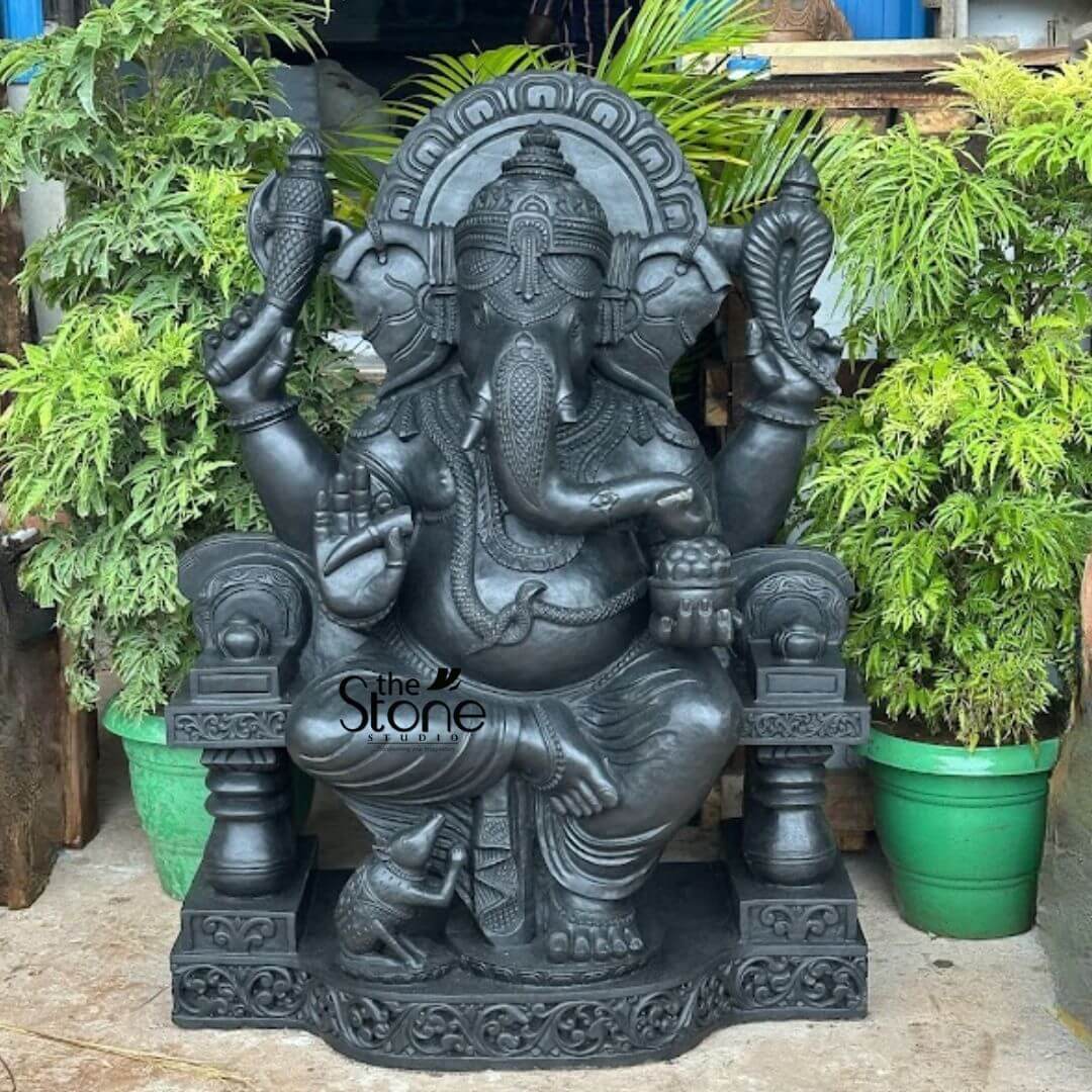 Black Stone Ganesha Sculpture for Garden 3.5ft 3 magnificent, large statue of Lord Ganesha, expertly carved from dark grey or black stone, showcased in a vibrant green outdoor setting. The detailed sculpture portrays Ganesha seated in a relaxed, regal posture with his characteristic elephant head, large ears, an elaborate crown, and a single broken tusk. His trunk gracefully curls to his left, touching a sweet modak held in his lower left hand. In his upper right hand, he wields an ankusha (goad), and in his upper left, a pasha (noose). His lower right hand is raised in the Abhaya mudra, a gesture of blessing and protection, while his lower left hand cradles a small bowl brimming with modaks. Ganesha's portly belly is adorned with intricate necklaces, armlets, bracelets, and a prominent waist belt shaped like a snake. He sits upon a beautifully carved throne with decorative pillars and a fan-shaped Prabhavali (archway of light) behind his head, all resting on a multi-tiered, ornate rectangular base featuring detailed scrollwork patterns. At his lower right foot, his loyal vahana, the small mouse Mushika, is depicted looking up at him. The statue exhibits exceptional craftsmanship with a smooth, matte finish, suggesting it's carved from a high-quality material like black granite or similar dark stone. The spiritual idol is framed by lush, vibrant green foliage from various potted plants on either side and behind, adding to its serene and natural aesthetic. A subtle 'The Stone Studio' watermark is visible on the statue's lower left side, indicating the artisan or supplier. This impressive and intricately detailed Ganesha statue embodies wisdom, prosperity, and the removal of obstacles, making it a perfect centerpiece for a grand garden, a dedicated puja room, or as an exquisite piece of art in a discerning home or temple environment.
