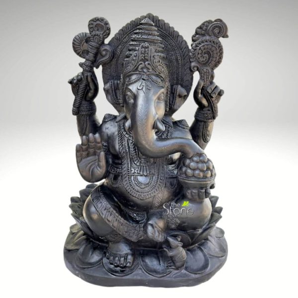 magnificent, dark, highly detailed stone sculpture of Lord Ganesha, the revered Hindu deity, is showcased against a plain, bright white background. The statue depicts Ganesha seated in a dignified Padmasana (lotus) position, symbolizing peace and meditation. His iconic elephant head features large, fanned ears, a gracefully curved trunk that reaches towards a bowl of sweets in his lower right hand, and a single tusk, symbolizing the ability to overcome duality. His small, discerning eyes convey wisdom and calm. Ganesha wears an elaborate, tiered crown (mukuta) adorned with intricate carvings, and a circular, finely sculpted 'prabhamandala' (aura or halo) frames his head, featuring traditional Indian motifs. He is depicted with four arms, symbolizing his divine power and ability to interact with the world on multiple levels. His upper left hand holds a ceremonial axe (parashu or ankusha), often associated with cutting through attachments and ignorance, while his upper right hand holds a noose (pasha), symbolizing the ability to capture desires or guide devotees. His lower left hand is raised in the Abhaya Mudra, a gesture of fearlessness, blessing, and protection, while his lower right hand lovingly holds a bowl overflowing with laddus (Indian sweet dumplings), from which his trunk is gracefully sampling a treat, representing material abundance and spiritual fulfillment. The deity has a prominent, pot-bellied form, symbolizing prosperity, generosity, and the absorption of the universe. He is adorned with multiple necklaces, armlets, and a decorated waistband, all intricately carved into the stone, highlighting the artisan's meticulous craftsmanship. His drapery is also richly detailed. The entire figure is seated upon a beautifully sculpted double-lotus pedestal (padma peetha), with each petal distinctly rendered. At his feet, a small, alert mouse (Mooshika), his traditional vahana (mount), is visible, perhaps reaching for a fallen laddu, symbolizing Ganesha's ability to control and guide all beings, even the smallest. Crafted from what appears to be polished black or very dark grey stone, possibly granite or a similar natural material, the sculpture exhibits a smooth finish in some areas and incredible textural depth in the carved details. The studio lighting emphasizes the fine artistry, casting subtle shadows that highlight the three-dimensional form and intricate patterns. This devotional sculpture embodies themes of wisdom, prosperity, new beginnings, and the removal of obstacles, making it a powerful visual for articles discussing Hindu deities, spiritual home decor, traditional Indian artistry, cultural symbolism, or the significance of Ganesha in spiritual practices. The natural stone finish adds to its timeless appeal and traditional authenticity.
