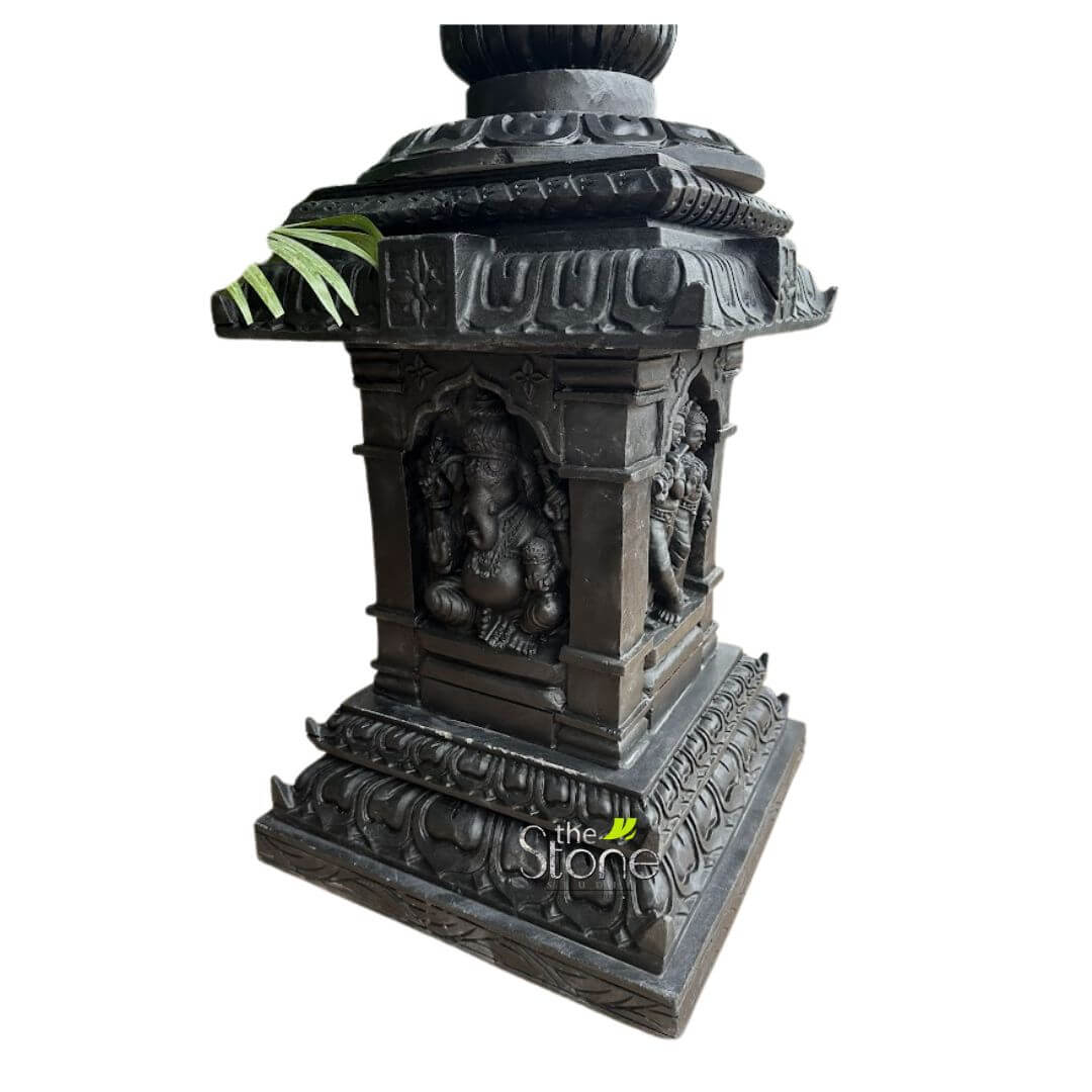 Granite Tulsi Pot 3ft 6 majestic, intricately hand-carved dark grey or black stone sculpture, resembling a traditional Indian temple pillar or a Deepa Stambha (lamp post) base, stands upright against a pure white background. The sculpture is highly detailed, showcasing profound craftsmanship. The central part of the structure is a rectangular body featuring deeply recessed niches on at least two visible sides. On the prominent front-facing side, a serene Lord Ganesha is depicted, seated comfortably in a relaxed posture within an elegant arched frame. He has his characteristic elephant head, four arms holding traditional attributes like a sweet modak and an axe, adorned with elaborate jewelry and a crown. His large belly and benevolent expression are clearly visible, conveying wisdom and prosperity. To the right of Ganesha, in a similar arched niche, another standing deity or figure is carved, though less distinctly visible, also adorned with traditional garments and possibly attributes. The sculpture's body is flanked by decorative pillars and topped with a multi-tiered, roof-like structure, reminiscent of classical Indian temple architecture with its intricate eaves and moldings. A single sprig of vibrant green leaves subtly emerges from under the eaves on the left side, adding a touch of natural contrast and suggesting the sculpture's potential placement in a lush garden, patio, or other outdoor spiritual setting. The base of the pillar is broad and multi-tiered, featuring exquisite traditional Indian design elements such as classical lotus petal motifs (Padma Pitha) and continuous scrolling foliage patterns, providing a stable and ornate foundation. The deep carvings create strong shadows and highlights, emphasizing the texture and three-dimensionality of the dark stone. This exquisite piece embodies rich Hindu iconography and traditional stone carving techniques, making it a significant representation of devotional art, garden decor, or a focal point for spiritual contemplation.
