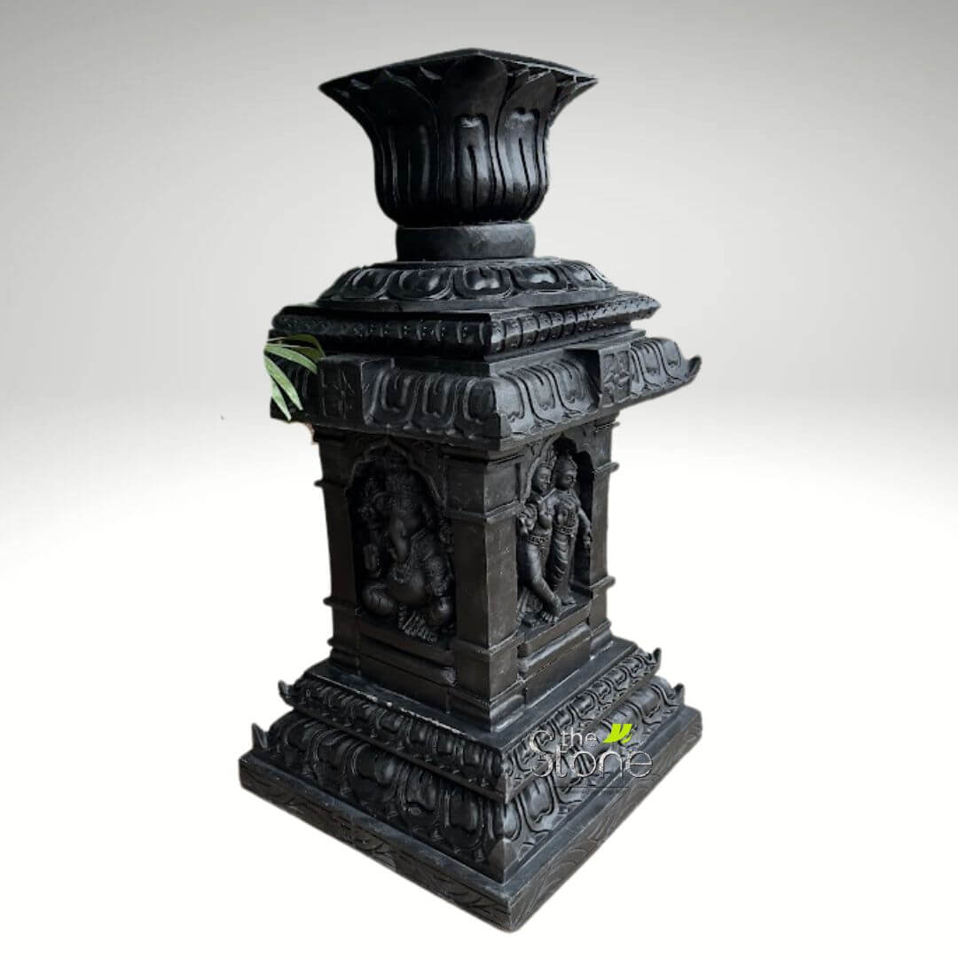 Granite Tulsi Pot 3ft 4 majestic and intricately carved dark gray sculptural pedestal, likely crafted from stone or a heavy resin, stands prominently against a soft, light gray background. The entire piece is rendered in a uniform, deep charcoal tone, which highlights the exquisite detail of its three-dimensional carvings without distracting colors. The pedestal is crowned with a prominent, fluted lotus-shaped finial or lamp holder, featuring deeply carved petals that open upwards, suggesting its use for holding a flame or flowers as a traditional "Deepastambha" (lamp tower) or "Vilakku Thoon" (lamp post) in South Indian temple architecture. Below this, the structure transitions into a square, multi-tiered capital adorned with continuous bands of ornate temple-style scrollwork and decorative motifs. The central square column forms the core, featuring arched niches on at least two visible sides, each housing a detailed carving of Hindu deities. On the left side, Lord Ganesha is depicted in a traditional seated `lalitasana` pose, his elephant head, benevolent expression, and prominent potbelly clearly visible. He appears to be holding his characteristic attributes, possibly a `modak` sweet and a broken tusk, rendered with fine detail. On the adjacent right side, an elegant divine couple, possibly `Radha-Krishna` or `Shiva-Parvati`, stands gracefully in intimate embrace or close proximity, adorned with traditional attire and jewelry. Their forms are rendered with fluid lines typical of classical Indian sculpture, framed within a similar arched niche. A small cluster of vibrant green leaves is subtly visible tucked into the left upper edge of the central column, providing a delicate natural contrast to the dark stone. The pedestal tapers outwards to a robust, multi-layered base, mirroring the intricate scrollwork and architectural elements of the upper sections, providing stability and grandeur to the entire piece. This finely crafted item exemplifies traditional Indian temple art and craftsmanship, serving as an ideal cultural home decor accent, a garden sculpture, or a devotional `murti` display stand. Its classic design and depiction of revered Hindu deities make it a significant and spiritual addition to `pooja rooms`, `meditation spaces`, or any setting appreciating `heritage sculpture` and `religious art`.