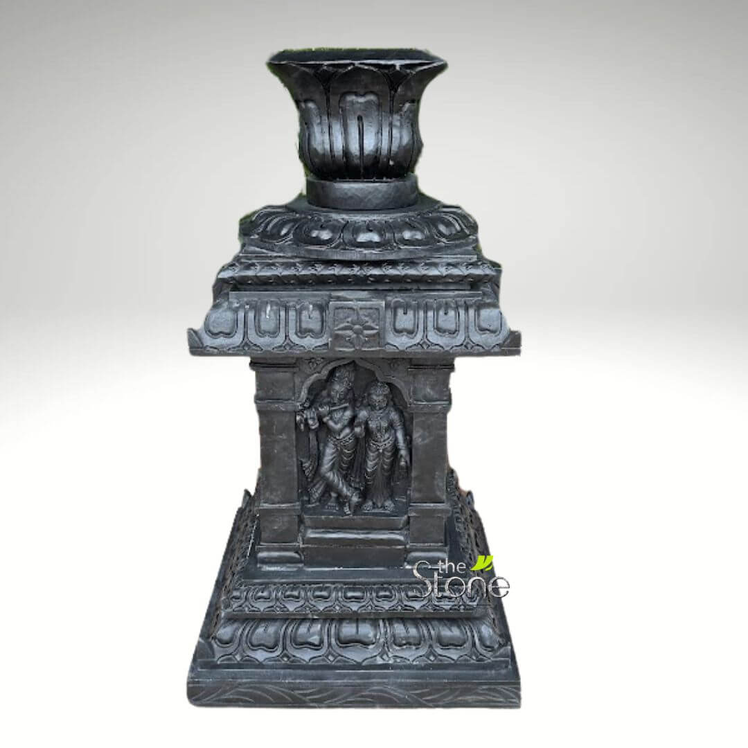 Granite Tulsi Pot 3ft 5 meticulously carved, dark black granite (or basalt) sculpture, designed as an ornate devotional shrine or decorative planter, is presented against a plain, light grey background. The sculpture stands tall and robust, showcasing intricate details typical of traditional Indian temple art. The uppermost section features a wide, fluted opening reminiscent of a blooming lotus flower or an antique urn, with pronounced vertical grooves and subtly scalloped edges. This suggests it could function as a holder for offerings like flowers, incense, or perhaps a small lamp (diya), connecting to its spiritual or decorative purpose. Below this elegant top, a multi-tiered structure forms the body of the sculpture. It comprises several carefully crafted cornices and friezes, adorned with elaborate architectural details and traditional Indian motifs, such as stylized lotus petals and intricate geometric patterns. These elements reflect the classical design principles found in Hindu temple architecture. The most prominent feature is a recessed central niche, which serves as a miniature shrine. It is beautifully framed by slender pilasters on either side, adding depth and architectural authenticity. Within this niche are exquisitely detailed carvings of the revered Hindu deities, Krishna and Radha. Krishna stands on the left, depicted in his iconic tribhanga (triple-bend) pose, holding a flute gracefully to his lips, his form adorned with delicate drapery, flowing garments, and intricate jewelry that signify his divine status. Radha stands gracefully beside him on the right, mirroring his intricate attire and ornamentation, her posture suggesting devotion and companionship. Both figures exude a serene and artistic beauty, capturing a moment of divine harmony. The sculpture rests on a broad, robust base, which is also layered with several ornate platforms. This base is decorated with continuous bands of meticulously carved lotus petal designs and subtle architectural mouldings, providing a stable foundation and adding to its grand and imposing appearance. This piece exemplifies the rich tradition of Indian stone carving and devotional art, combining profound spiritual symbolism with masterful craftsmanship. Its dark, polished surface, likely black granite or a similar dense stone, enhances the depth and contrast of the intricate carvings, making it a striking representation of Hindu cultural heritage and the divine love embodied by Krishna and Radha. It serves as an ideal accent for spiritual spaces, traditional decor, or as a focal point in a curated collection focusing on Indian art and spirituality.
