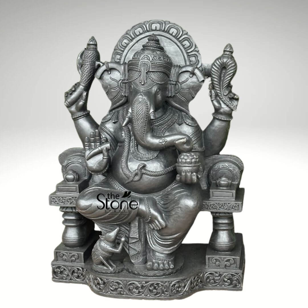 Black Stone Ganesha Sculpture for Garden 3.5ft 4 magnificent and intricately carved statue of the Hindu deity Ganesha, crafted from a dark grey or black stone, possessing a smooth, lustrous finish that highlights its fine details. Ganesha is depicted seated in a regal posture, likely Lalitasana, with his right leg resting comfortably on the pedestal while his left leg is folded, conveying a sense of serene authority. His iconic elephant head features large, gracefully carved ears, a prominently curled trunk pointing towards a bowl of sweets in his lower left hand, and a single intact tusk, signifying his wisdom and ability to overcome duality. A traditional mukuta (crown) adorns his head, intricately detailed with patterns, and a subtle third eye is visible on his forehead. His portly belly is adorned with a nagabandha (snake belt), symbolizing cosmic energy. He wears various ornate jewelry, including necklaces, armlets, bracelets, and anklets, all meticulously carved with traditional designs. With four arms, Ganesha holds his characteristic attributes: in his upper right hand, he grasps an ankusha (elephant goad), and in his upper left, a pasha (noose), representing his ability to guide and control. His lower right hand is raised in the Varada Mudra, a gesture of blessing and granting boons, with the palm facing forward and fingers pointing downwards. His lower left hand holds a bowl filled with modaks (sweet dumplings), a treat he is famously fond of. He sits upon a grand, elaborately carved throne featuring ornate pillars, armrests, and a majestic circular prabhamandala (aura or halo) behind his head, signifying his divine stature. The multi-tiered pedestal beneath the throne is richly adorned with intricate traditional Hindu patterns and scrollwork. His faithful vahana, Mushika (the mouse), is positioned attentively at his feet on the pedestal, appearing to look up at him or perhaps towards the sweets. The entire statue is presented against a plain, light grey background, ensuring full focus on its detailed craftsmanship. The watermark "the Stone Studio" is visible on the lower left, indicating the source of this exquisite stone sculpture or idol. This stunning Ganesha statue, perfect for home decor, spiritual practices, or as a focal point in a sacred space, exemplifies traditional Indian stone carving artistry and connects to themes of spirituality, remover of obstacles, wisdom, and prosperity often associated with Lord Ganesha.