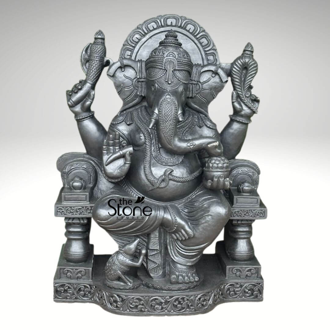 Black Stone Ganesha Sculpture for Garden 3.5ft 1 majestic and intricately detailed dark gray stone statue of Lord Ganesha, presented by "The Stone Studio," sits prominently against a neutral, light gray background. The revered Hindu deity, known for removing obstacles, is depicted seated on an ornate, high-backed throne or simhasana. His posture is relaxed yet regal, with his left leg folded in a meditative padmasana and his right foot resting gracefully on the elaborately carved pedestal below. Ganesha's iconic elephant head features large, expressive ears, a prominent trunk curled gently to his left, and a single tusk, symbolizing wisdom and sacrifice. His portly belly, a symbol of prosperity and generosity, is adorned with an intricate cobra necklace, while various other traditional ornaments like a detailed crown (mukut), armlets, wristlets, and ankle bracelets are meticulously sculpted onto his form. He possesses four arms, each holding symbolic objects: the upper left hand holds an ankusha (goad), the upper right hand holds what appears to be a conch shell or a patterned palm leaf. His lower right hand cradles a small bowl brimming with modaks (sweet dumplings), and his lower left hand is raised in the protective Abhaya Mudra, offering blessings and reassurance. At his feet, his loyal vahana, Mushika (the mouse), kneels reverently, gazing up at the deity, seemingly ready to partake in the modaks. The throne itself is richly carved with elaborate patterns, including an arched prabhamandala (aureole) behind Ganesha's head and delicate scrollwork along the base and supporting pillars, highlighting the fine craftsmanship. The entire sculpture boasts a consistent, elegant dark charcoal gray finish, suggesting it's expertly crafted from high-quality stone like granite or basalt, giving it a timeless and substantial presence. This exquisite piece embodies traditional Indian artistry and devotion, making it ideal for spiritual decor, home altars, or as a striking focal point in any space, reflecting the quality and aesthetic offered by "The Stone Studio."