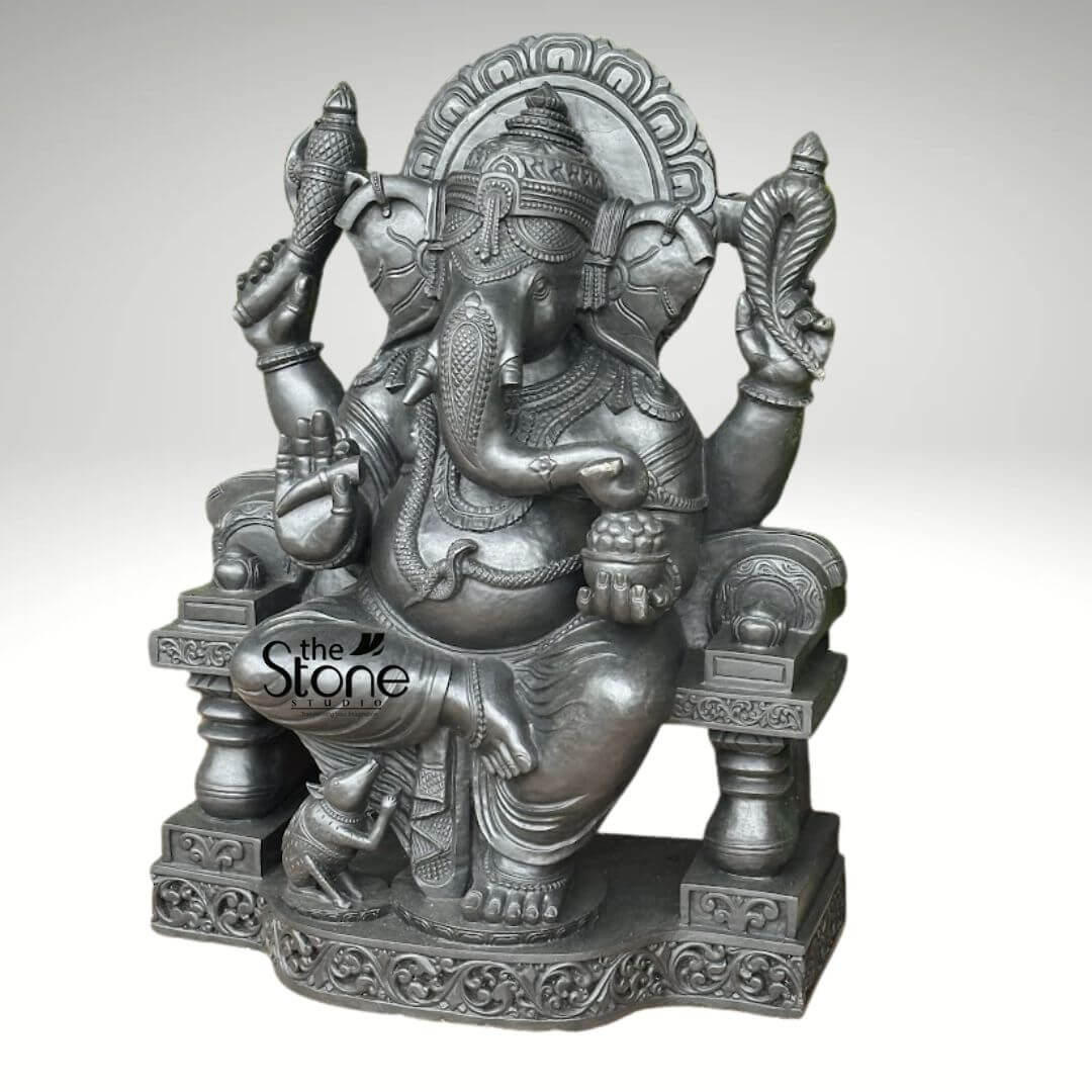 Black Stone Ganesha Sculpture for Garden 3.5ft 2 highly detailed and majestic dark grey stone sculpture of Lord Ganesha, the revered Hindu deity, seated regally on an intricately carved throne. His iconic elephant head features prominent ears, a gently curving trunk turned towards his left, and one intact tusk, while the other is characteristically absent. He wears an elaborate crown and intricate jewelry, including necklaces, armlets, and a waist belt, with a draped garment around his legs. With four arms, a sign of his divine power, Ganesha holds various symbolic objects. His upper right hand clasps a ritual implement or perhaps his broken tusk (danta), while his lower right hand is raised in the Abhaya Mudra, a gesture of blessing and reassurance, signifying protection and fearlessness. His upper left hand holds an ankusha (elephant goad), representing his ability to guide devotees and remove obstacles, and his lower left hand cradles a bowl brimming with modaks, his favorite sweet, symbolizing the rewards of spiritual pursuit and the sweetness of liberation. He rests upon a ceremonial throne, adorned with elaborate carvings on its base and columns. Behind his head, an ornate prabhamandala (halo) with intricate scrollwork further emphasizes his divine presence and wisdom. At his left foot, his loyal vahana, the tiny mouse (Mushika), sits attentively, looking up at his lord, symbolizing humility and the power of the smallest creatures. The sculpture possesses a smooth, dark metallic grey finish, suggesting it is crafted from a durable material like granite, basalt, or a high-quality composite stone, emphasizing its robust and timeless quality. The 'The Stone Studio' watermark on the lower left reinforces its craftsmanship as a premium stone product. This exquisite piece, embodying spiritual significance and artistic mastery, is ideal for enhancing sacred spaces, home altars, garden landscapes, or as a focal point in a collection of traditional Indian art, making it a perfect visual complement for articles discussing Hindu mythology, deity symbolism, stone craftsmanship, or spiritual home decor.