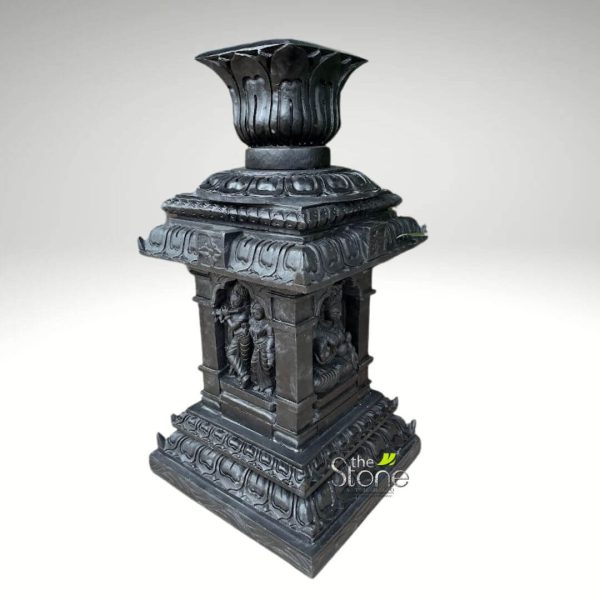 majestic, intricately carved dark grey stone pillar or Deepastambham (traditional lamp post base) stands prominently against a plain, light grey studio background. The sculpture is rendered in a traditional South Indian temple style, showcasing exceptional craftsmanship and rich detail. Crowning the structure is an elegant, multi-layered lotus finial, its petals deeply sculpted and pointing upwards, designed to potentially hold a lamp or serve as a decorative element. Below this, a series of progressively larger, tiered square sections form the capital, each adorned with stylized lotus petal patterns and geometric borders, characteristic of classical Indian architecture. The central, square shaft of the pillar is the most ornate, featuring recessed arched niches on at least two visible sides. On the left, a devotional scene unfolds: the Hindu deity Krishna, identifiable by his flute, stands gracefully in a dynamic pose, accompanied by Radha or a Gopi, both figures rendered with delicate drapery, traditional attire, and serene expressions that evoke spiritual connection. On the adjacent visible side, a multi-armed Hindu deity is depicted seated in a meditative or royal posture, signifying divine power and tranquility. This figure, possibly Vishnu given the nearby Krishna, is sculpted with intricate details, including ornate headdresses and jewelry, typical of ancient Indian iconography. The robust base is also multi-tiered and square, echoing the decorative motifs of the upper sections with repeating floral or architectural patterns, providing a solid and aesthetically balanced foundation. The entire piece exudes a sense of traditional craftsmanship and sacred symbolism. The dark hue, resembling polished black granite or a dark basalt stone, enhances the depth and intricacy of the carvings, giving it an antique and timeless appeal. This elaborate stone sculpture serves as an excellent visual representation for articles discussing traditional Indian stone carving, Hindu religious art, South Indian temple design, spiritual garden decor, or bespoke architectural elements that blend cultural heritage with aesthetic beauty. Its presence evokes a sense of tradition, devotion, and masterful artistry, making it a significant cultural artifact or a striking piece of decor.