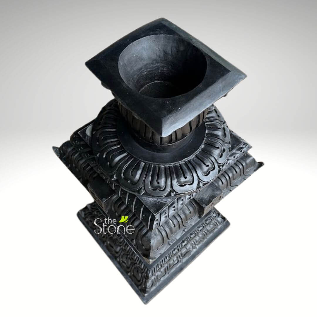 Granite Tulsi Pot 3ft 3 stately, dark black hand-carved stone pedestal or plinth, presented against a clean, light grey to white studio background. The sculpture is viewed from a slightly elevated, angled perspective, showcasing its multi-tiered, square-based structure and intricate detailing. At the very top, a prominent, smooth, square platform features a deep, circular concave depression, suggesting its use as a holder for a lamp, a candle, an incense burner, a small plant, or offerings. The edges of this top platform are sharply defined. Below the top, the piece transitions into multiple square tiers, each meticulously carved and progressively smaller as they ascend, creating a pyramidal effect. The most striking decorative element is a repeating pattern of stylized lotus petals, intricately carved in relief around a prominent circular band on the upper-middle tiers. Further down the body of the pedestal, additional friezes display intricate geometric motifs and traditional architectural patterns, adding layers of texture and visual interest to the dark stone surface. The deep carvings create shadows that highlight the craftsmanship and depth of the design. The robust, solid square base provides a strong foundation for the entire structure, which appears to be crafted from heavy, durable black stone or granite, with a subtly textured, matte finish. The overall design suggests a traditional or spiritual aesthetic, making it suitable as a decorative sculpture, an architectural element, a garden ornament, a planter base, or a stand for religious or ceremonial items. The logo "the Stone STUDIO" is subtly visible in the lower left quadrant, indicating the artisanal origin and specialized nature of this unique piece of stone art.