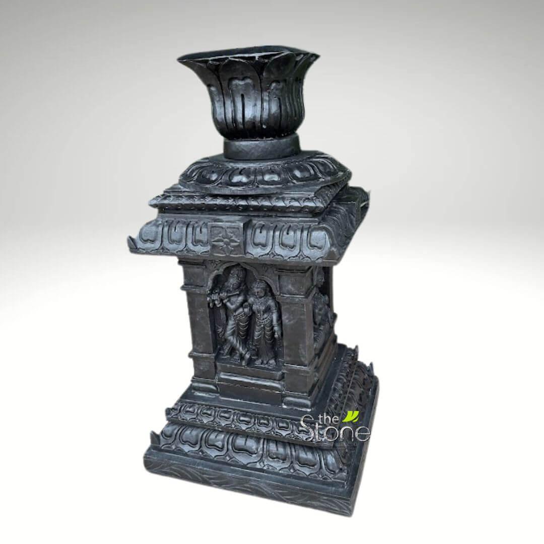 Granite Tulsi Pot 3ft 2 meticulously carved, dark grey stone sculpture, likely a decorative lamp stand or architectural element, stands prominently against a plain, soft grey background. This intricate piece exemplifies traditional Indian temple craftsmanship, possibly made from a dense material like granite or basalt. The sculpture features a multi-tiered structure, beginning with a substantial, stepped square base. This base is richly adorned with repeating, deeply carved lotus petal motifs along its edges, signifying purity and beauty in Hindu iconography. Rising from this foundation is the main body, a square pavilion-like structure. Each visible side of this central section showcases a recessed niche or alcove. On the front-facing niche, two meticulously detailed figures are carved in bas-relief. The taller figure, likely representing Lord Krishna, is depicted gracefully holding a flute to his lips. Beside him stands another figure, possibly Radha, adorned in traditional attire and jewelry. Both figures are rendered with fine detail, capturing their divine attributes and traditional poses, set within miniature architectural frames of pillars and archways, reminiscent of a small temple shrine. Partially visible on the right side, another niche appears to house similar divine or mythological figures, suggesting a continuous narrative or theme around the piece. Above the central pavilion, the sculpture transitions into several stacked, receding square tiers, each decorated with continuous patterns and subtle overhangs, reflecting the architectural style of a temple's vimana or gopuram. The entire piece culminates in an elegant, inverted lotus bud-shaped finial, deeply fluted with petal-like grooves, designed either to hold an oil lamp or simply serve as a decorative crowning element. The overall texture appears matte, highlighting the detailed chisel work and the natural grain of the stone. A subtle watermark in the lower right corner reads "the Stone" with a stylized leaf emblem, emphasizing the natural material and artisanal origin of this cultural and decorative artwork. This stone carving is a testament to traditional Indian art forms, perfect for enhancing home decor, spiritual spaces, or as a collector's item reflecting rich cultural heritage.
