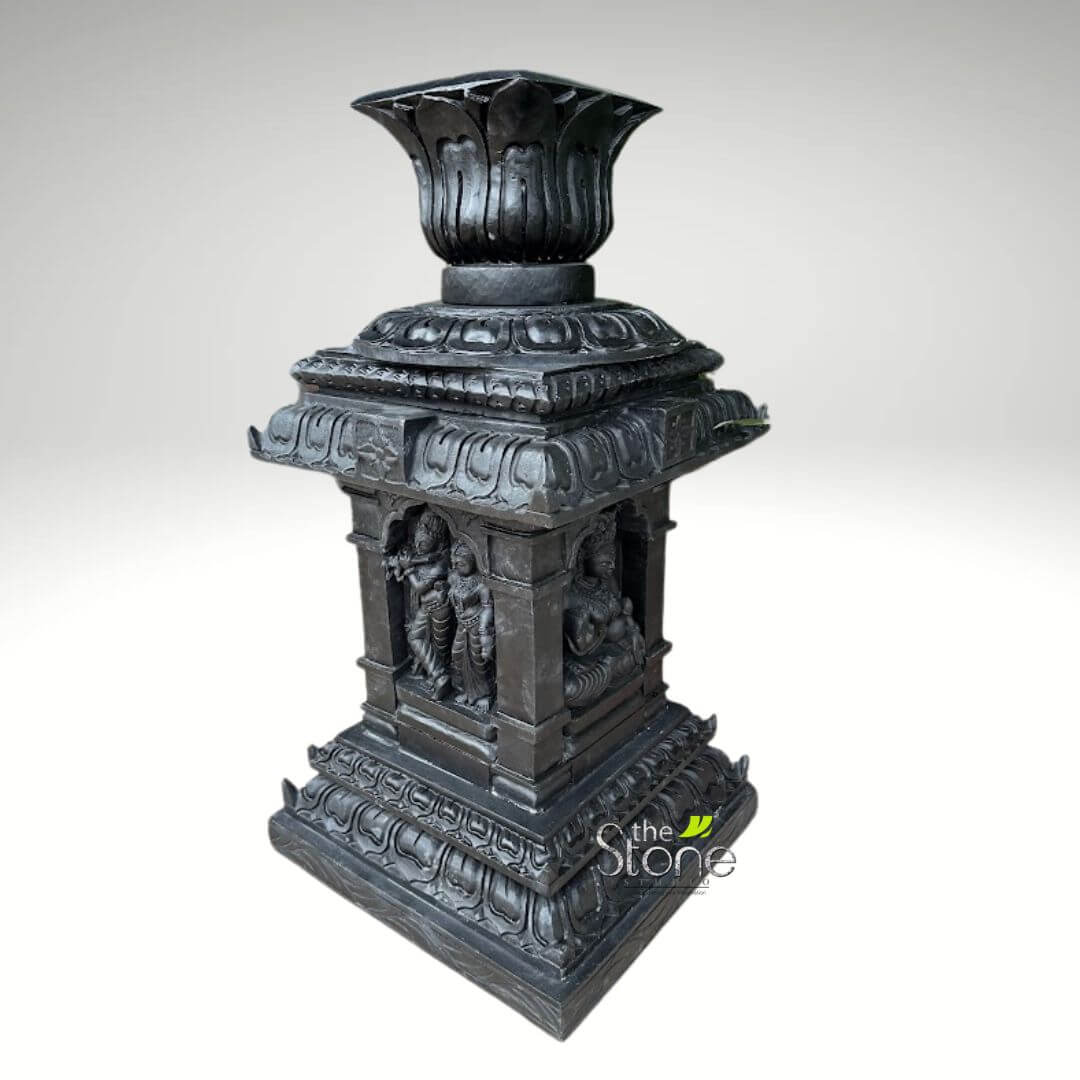 Granite Tulsi Pot 3ft 1 majestic, intricately carved dark grey stone pillar or Deepastambham (traditional lamp post base) stands prominently against a plain, light grey studio background. The sculpture is rendered in a traditional South Indian temple style, showcasing exceptional craftsmanship and rich detail. Crowning the structure is an elegant, multi-layered lotus finial, its petals deeply sculpted and pointing upwards, designed to potentially hold a lamp or serve as a decorative element. Below this, a series of progressively larger, tiered square sections form the capital, each adorned with stylized lotus petal patterns and geometric borders, characteristic of classical Indian architecture. The central, square shaft of the pillar is the most ornate, featuring recessed arched niches on at least two visible sides. On the left, a devotional scene unfolds: the Hindu deity Krishna, identifiable by his flute, stands gracefully in a dynamic pose, accompanied by Radha or a Gopi, both figures rendered with delicate drapery, traditional attire, and serene expressions that evoke spiritual connection. On the adjacent visible side, a multi-armed Hindu deity is depicted seated in a meditative or royal posture, signifying divine power and tranquility. This figure, possibly Vishnu given the nearby Krishna, is sculpted with intricate details, including ornate headdresses and jewelry, typical of ancient Indian iconography. The robust base is also multi-tiered and square, echoing the decorative motifs of the upper sections with repeating floral or architectural patterns, providing a solid and aesthetically balanced foundation. The entire piece exudes a sense of traditional craftsmanship and sacred symbolism. The dark hue, resembling polished black granite or a dark basalt stone, enhances the depth and intricacy of the carvings, giving it an antique and timeless appeal. This elaborate stone sculpture serves as an excellent visual representation for articles discussing traditional Indian stone carving, Hindu religious art, South Indian temple design, spiritual garden decor, or bespoke architectural elements that blend cultural heritage with aesthetic beauty. Its presence evokes a sense of tradition, devotion, and masterful artistry, making it a significant cultural artifact or a striking piece of decor.