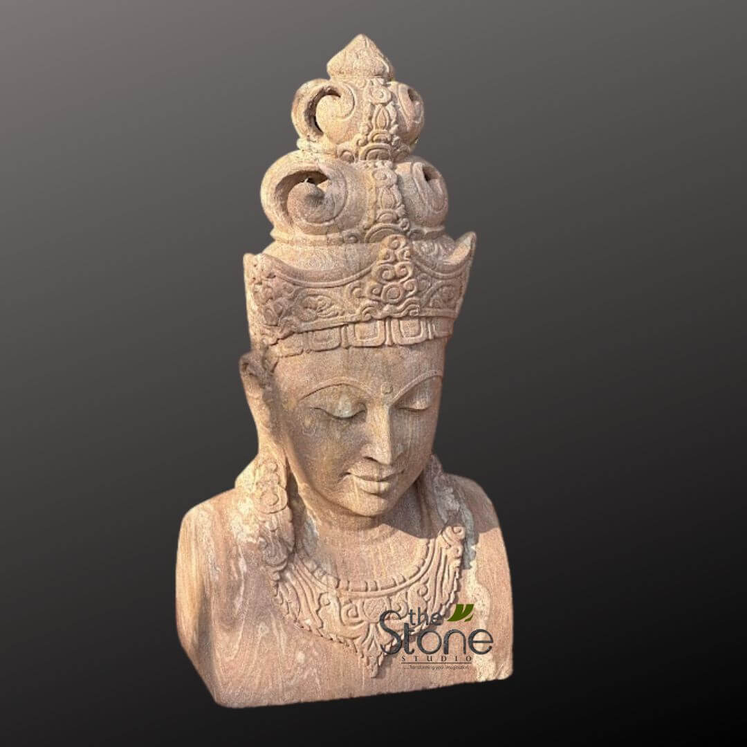 2ft Lord Vishnu Head 4 highly detailed, hand-carved stone sculpture, likely a bust, depicts the head and upper torso of a serene deity or enlightened being, possibly a Buddha or Bodhisattva. The sculpture is crafted from a light brown or reddish-beige sandstone, exhibiting a distinct natural grain and a slightly porous texture that emphasizes its organic material and hand-finished quality. The figure has a calm, meditative expression, with eyes gently closed and downcast, and a subtle, peaceful smile on its lips, conveying a sense of inner tranquility and wisdom. An elaborate, multi-tiered crown adorns the head, featuring intricate traditional Asian patterns, spirals, and geometric designs, culminating in a prominent, pointed finial at its peak. Large, decorative earrings hang from the elongated earlobes, and an ornate necklace with complex floral and geometric carvings gracefully drapes around the neck and upper chest area. The craftsmanship is exquisite, showcasing the fine details in the facial features, the delicate lines of the jewelry, and the rich ornamentation of the headdress. The entire piece reflects skilled artisan work, highlighting the beauty and spiritual depth often found in ancient or traditional stone art. The sculpture is presented against a plain, dark grey, almost black background, which makes its earthy tones and intricate carvings stand out prominently. A subtle watermark for "The Stone Studio" is visible in the lower right, indicating its origin or creator, suggesting the image is likely showcasing the studio's expertise in crafting spiritual, artistic, and decorative stone pieces perfect for home decor, garden aesthetics, or as a focal point in a tranquil setting.