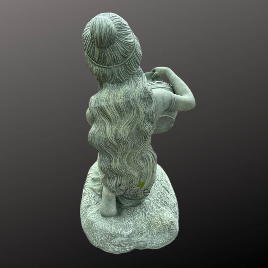 Sitting Lady Water Fountain 3ft 6 beautifully detailed natural stone sculpture, viewed from a slightly elevated angle looking over the figure's right shoulder, features a gracefully posed female figure. She is depicted kneeling or sitting on an organically shaped, textured rock formation that serves as her base. Her long, intricately sculpted wavy hair cascades down her back, with an elaborate bun or updo adorned by a band at the crown. Both arms are engaged with a classical-style round water pot or vessel; her left arm cradles it securely against her side, while her right hand delicately rests on or within its opening, suggesting an act of pouring, tending, or carrying water. The garden statue exhibits a captivating, weathered greenish-grey patina, reminiscent of aged natural stone, verdigris on bronze, or moss-covered rock, which enhances its timeless and antique aesthetic. The rough, artisanal texture of the stone is clearly visible in the details of her flowing locks, the robust form of the water jar, and the naturalistic ruggedness of the base. This high-quality carving exemplifies skilled craftsmanship and attention to detail, making it a focal point for any setting. The figure stands out prominently against a plain, dark grey to black gradient background, which serves to highlight the sculpture's form, texture, and muted coloration without distraction. This elegant piece of outdoor decor, possibly from "We're Stone" as indicated by the faint watermark, would be an ideal garden accent, a focal point for a patio or courtyard, or an elegant element in a water feature, adding a touch of classical artistry and tranquility to any landscape design or architectural space.