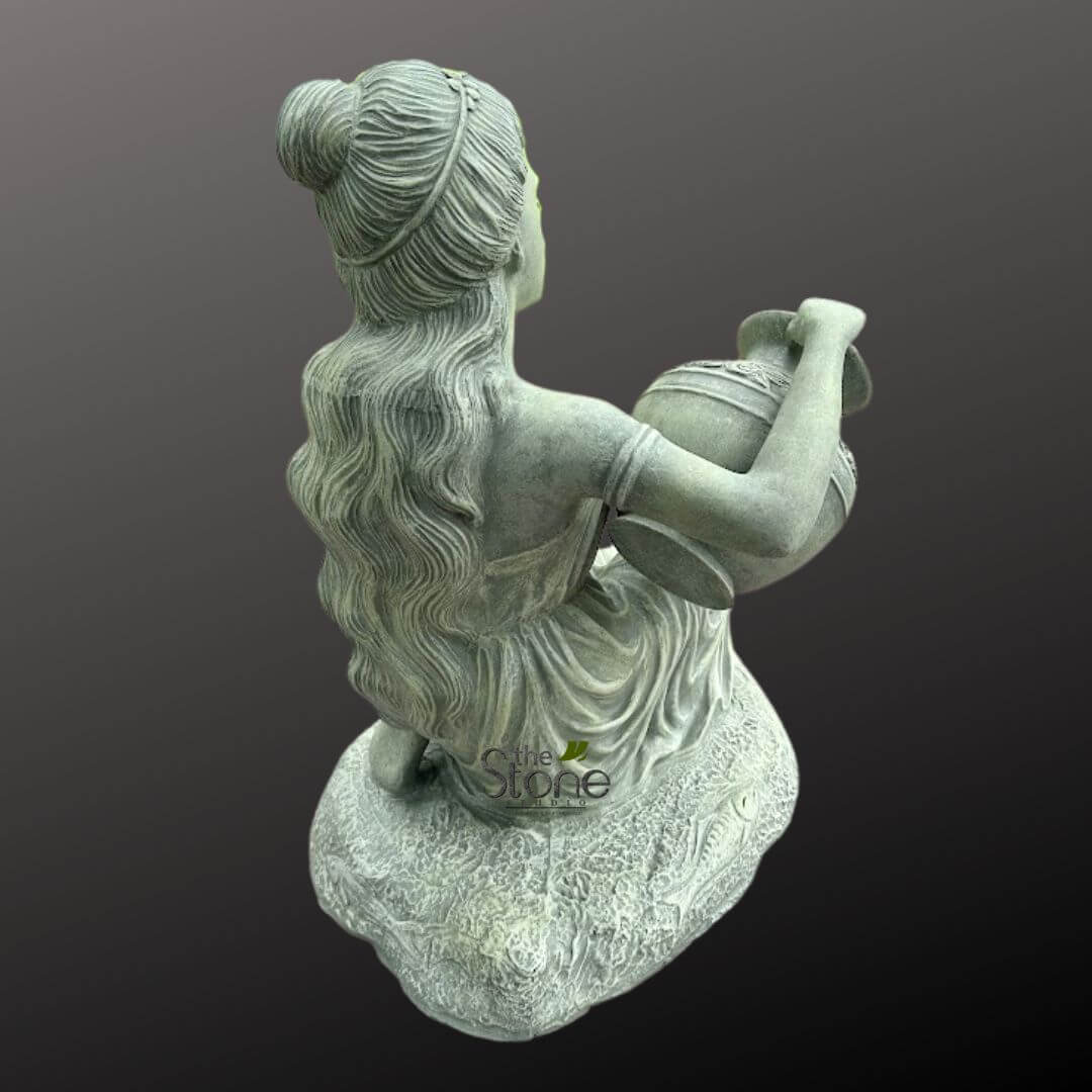 Sitting Lady Water Fountain 3ft 4 beautiful, detailed hand-carved stone sculpture of a graceful woman, presented from a slightly elevated, three-quarter rear view. The statue, likely intended as a garden ornament or decorative outdoor decor, depicts a figure kneeling or seated, with her body gently turned to her left, looking slightly upwards. Her long, flowing, intricately carved wavy hair cascades down her back, styled with a neat bun at the crown and a subtle headband, characteristic of traditional Indian sculptures or classical figures. She wears a flowing, draped garment, indicated by numerous soft folds and pleats. In her arms, she cradles a highly decorative kalash or water pot, adorned with ornate banded patterns in relief around its circumference and neck. The entire figurine is rendered in a verdant grey stone, possibly granite or limestone, exhibiting a rich, weathered patina that enhances the intricate details and gives it an aged, authentic look. She rests upon a textured, organically shaped base, which is subtly carved with a delicate, stylized floral or abstract pattern, suggesting a woven mat or natural rock formation. The plain, dark background draws full attention to the exquisite craftsmanship of this artisan-made piece. The watermark "The Stone Studio" is visible on the lower front of the base, identifying the creator or vendor of this magnificent figurine, which perfectly exemplifies high-quality stone artistry for home and garden design.