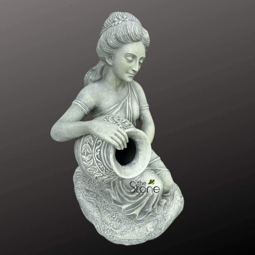 Sitting Lady Water Fountain 3ft 3 detailed, light grey stone sculpture depicts a serene female figure seated gracefully, looking downwards with a contemplative expression. Her hair is elegantly styled in a braided bun, and she wears a flowing, draped garment reminiscent of a classical or traditional Indian dress, with subtle bangles adorning her bare forearms. In her lap, she holds a beautifully ornamented, rotund pot or urn, tilted slightly forward. The pot features intricate relief carvings of lush leaves and swirling floral patterns, adding to its artistic appeal. The woman sits on a sculpted base that mimics draped fabric, which also has subtle relief designs. The entire sculpture has a weathered, antique stone or concrete finish, characterized by lighter grey highlights and darker shadows in the crevices, expertly enhancing the textures and intricate details of the carving. This gives the piece a timeless, aged appearance. Presented against a plain, dark grey background, the focus remains entirely on the craftsmanship and artistry of the sculpture. A subtle 'The Stone Studio' watermark is visible on the pot. This elegant and classical stone art piece would serve as a tranquil focal point for garden decor, a sophisticated patio accent, or an indoor statement piece, reflecting traditional aesthetics and a sense of calm. It's ideal for those seeking handcrafted stone art, classical garden statues, or a unique water feature component for their home and garden.