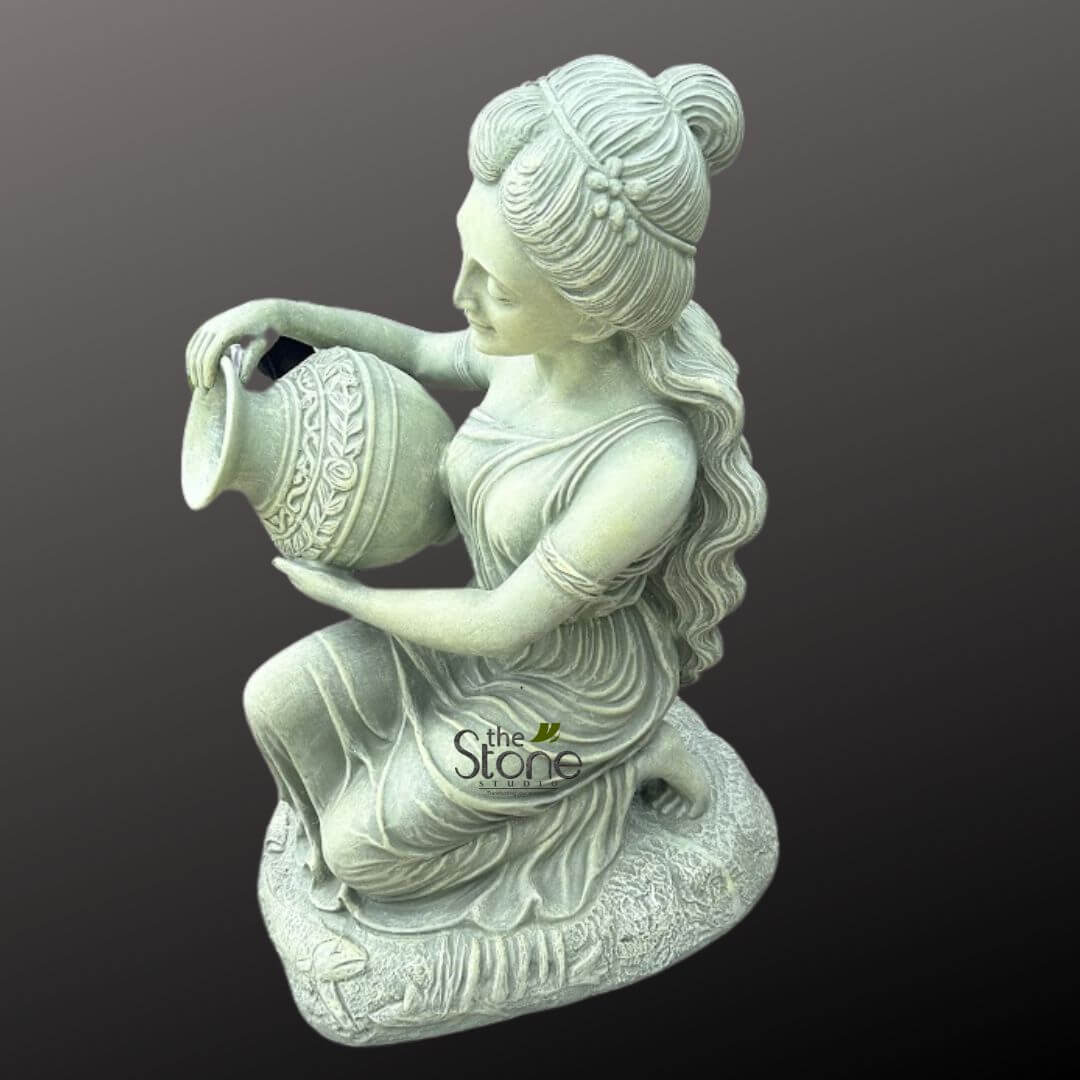 Sitting Lady Water Fountain 3ft 2 long, descriptive alt text for the image: "A detailed stone sculpture of a serene woman, likely crafted from light grey-green weathered concrete or cast stone, is depicted kneeling gracefully and appearing to pour water from an ornate, classical urn held in both hands. The woman, viewed from a slightly elevated side angle against a plain dark grey background, is dressed in a flowing, classical-style gown with visible drapes and natural folds that convey movement. Her hair is elegantly styled in an elaborate bun atop her head, with long, wavy strands cascading down her back, and a delicate floral embellishment adorns her hair near her temple. She has a gentle, downward gaze directed towards the decorative urn, which is beautifully textured and adorned with intricate carved bands of floral and leaf patterns around its circumference. The woman is positioned on a natural-looking, textured base, resembling a sculpted rock formation or patch of ground, which grounds the sculpture. This elegant and timeless garden ornament, with its fine craftsmanship and classical aesthetic, is an ideal piece for enhancing outdoor spaces like gardens, patios, or entryways, possibly functioning as a tranquil fountain or a standalone decorative art piece. The image highlights the detailed artistry and durable quality of handcrafted stone decor."