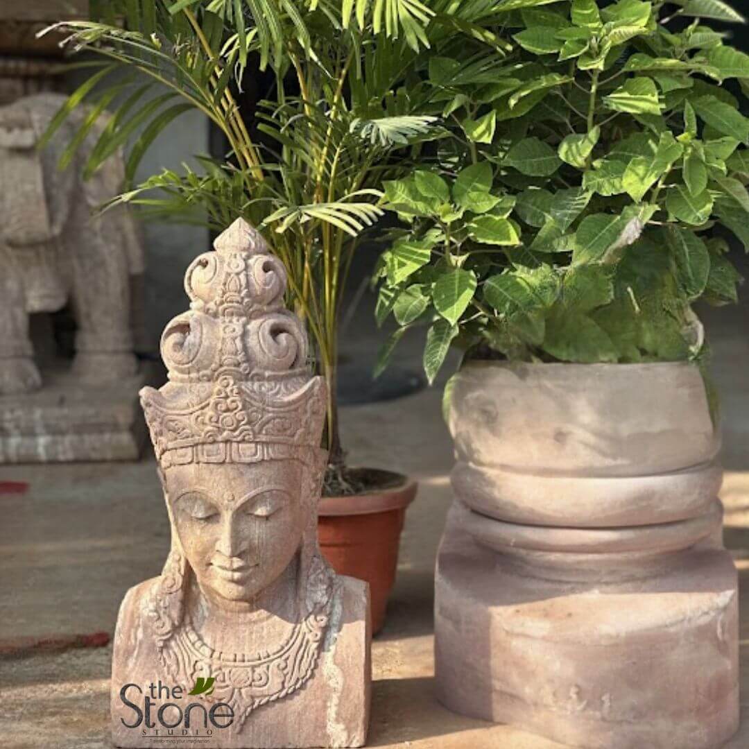 2ft Lord Vishnu Head 2 detailed view of handcrafted natural stone art and garden decor, featuring a serene deity bust and a large planter. In the foreground on the left, a striking, intricately carved stone sculpture of a bust from the chest up captures attention. Made from light reddish-brown natural sandstone, the figure, possibly a Bodhisattva or similar spiritual entity, exhibits a peaceful expression with closed eyes, soft lips, and elongated earlobes. An elaborate, multi-tiered crown or headdress adorns the head, showcasing delicate, swirling, traditional patterns. Below the face, a detailed necklace or garment collar with intricate designs is visible. The texture of the stone shows subtle variations and craftsmanship. To the right, a substantial, cylindrical stone planter complements the sculpture, appearing to be crafted from the same earthy-toned natural stone. Its design features a solid base with a wider, rounded section above, subtly tapering upwards, creating an organic, stacked look. The planter holds a lush, vibrant green leafy plant, possibly a variety of ficus or a similar shrub, with its dense foliage catching natural light. In the background, behind the stone bust, a tall, slender palm plant in a simple reddish-brown terracotta pot adds another layer of greenery. On the far left, a partial glimpse of another large stone carving, possibly an elephant statue, is visible, reinforcing the theme of extensive stone artistry. The entire scene is set outdoors, likely a garden or patio display area, bathed in natural light that highlights the textures and artisanal quality of the stone pieces. The "The Stone Studio" logo, featuring a stylized leaf, is subtly placed in the bottom left corner, indicating the origin of these handcrafted stone art and garden decor items. This image effectively showcases elegant stone craftsmanship, spiritual art, and landscaping elements designed to enhance tranquil outdoor or indoor spaces.