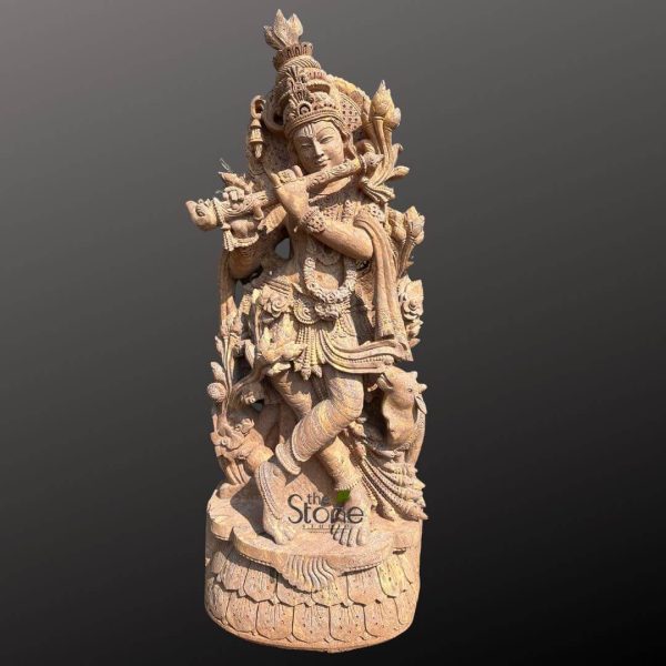 magnificent, intricately carved sandstone statue of Lord Krishna, depicted in his iconic tribhanga (triple-bend) posture, gracefully stands with his left leg crossed over his right. The light brown, granular texture of the natural sandstone lends a warm, earthy tone to the entire sculpture, highlighting the incredible depth and precision of the traditional Indian carving techniques. Krishna is shown holding a bansuri (bamboo flute) horizontally to his lips, poised as if playing a divine melody. His serene face is framed by an elaborate mukuta (crown) featuring multiple tiers of ornate carving and a prominent finial, possibly adorned with a subtle peacock feather motif. His forehead bears a tilak, and his ears are adorned with heavy, detailed earrings. His bare torso is draped with multiple necklaces, garlands, and armbands, showcasing the traditional ornamentation of a Hindu deity. A gracefully flowing uttariya (upper cloth) or dupatta wraps around his shoulders, adding to the dynamic flow of the sculpture. His lower body is clad in a richly detailed dhoti with intricate pleats and designs, secured by an ornate waist belt with hanging embellishments. Behind him rises a beautifully sculpted prabhamandala (aura or archway), overflowing with delicate floral patterns, foliage, and subtle symbolic elements, all carved from the same stone, creating a stunning backdrop. The entire sculpture rests on a multi-layered, round lotus pedestal (padma-pitha), meticulously carved with petaled details, grounding the divine figure. The statue is presented against a plain, dark gray background, emphasizing its artistic details and making it a striking focal point. A watermark "the Stone Hill" is visible on the lower front, indicating the source or craftsmanship of this detailed stone art piece, suggesting its relevance for articles on spiritual decor, Hindu sculpture, Indian craftsmanship, or large-scale deity statues.